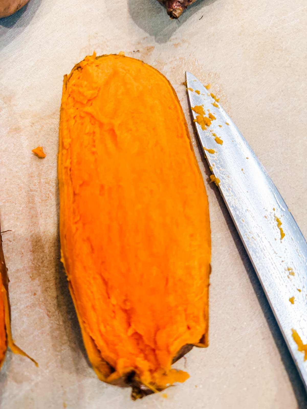 A cooked sweet potato sliced in half lengthwise sits on a light surface next to a knife with orange residue on the blade.
