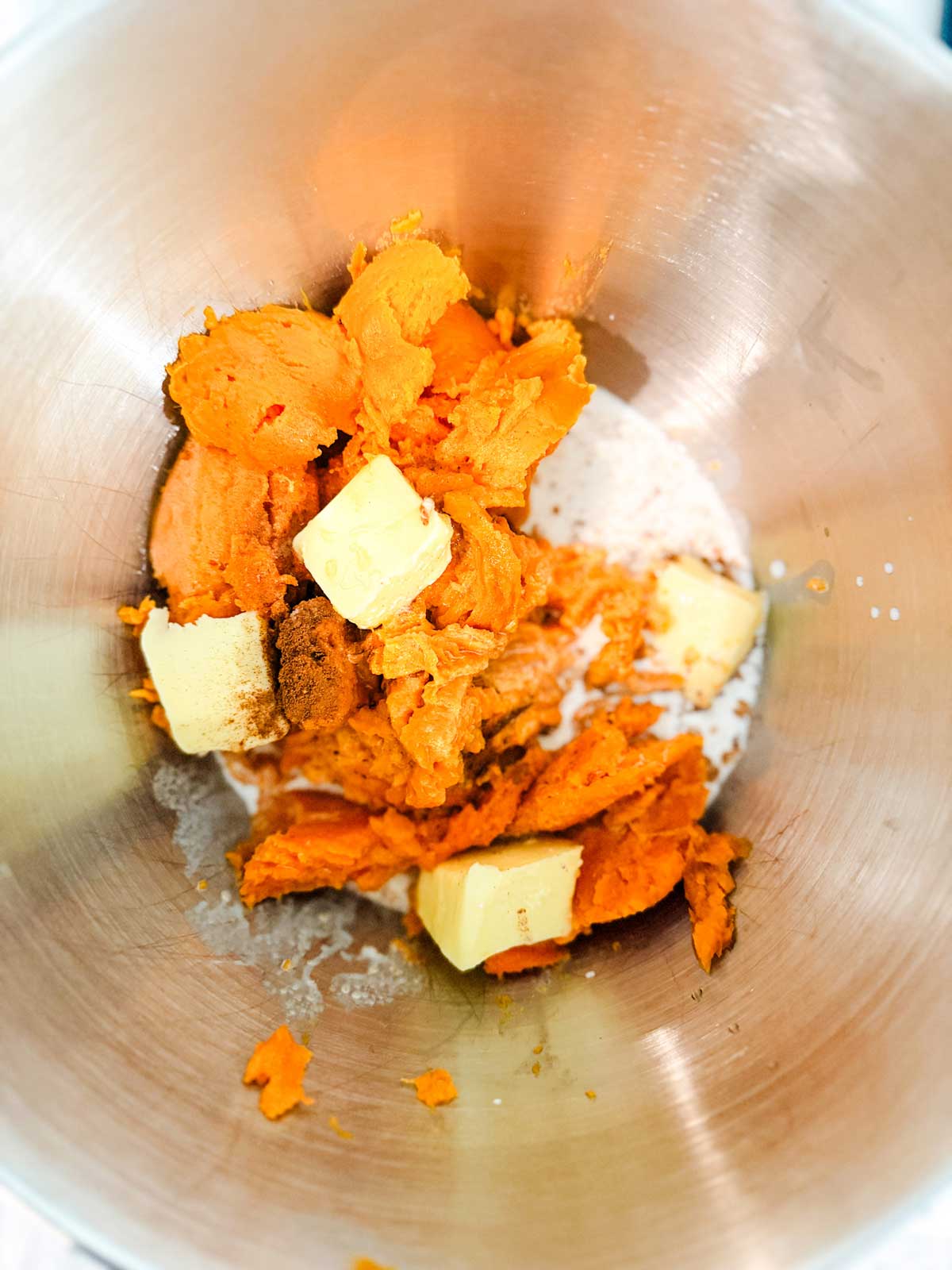 A mixing bowl containing scoops of mashed sweet potatoes, chunks of butter, sugar, and spices, ready to be mixed together.