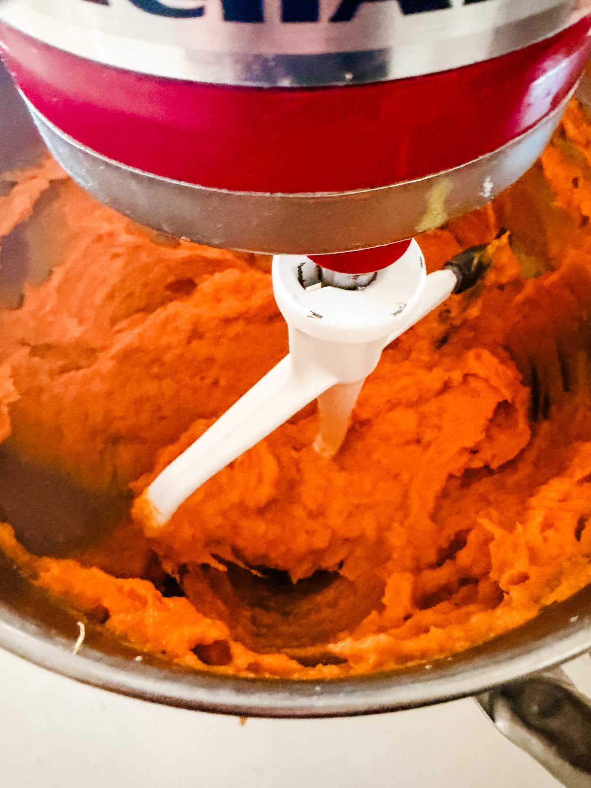 A close-up of a stand mixer blending bright orange mashed sweet potatoes in a stainless steel mixing bowl.