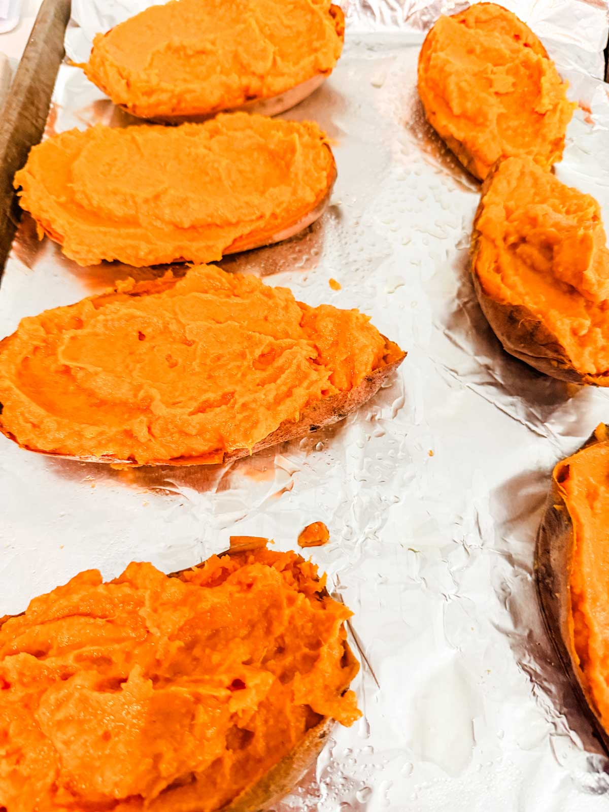 Six baked sweet potato halves with mashed filling are arranged on a foil-lined baking sheet, ready for the next step in preparation. The orange filling contrasts with the shiny silver of the foil.