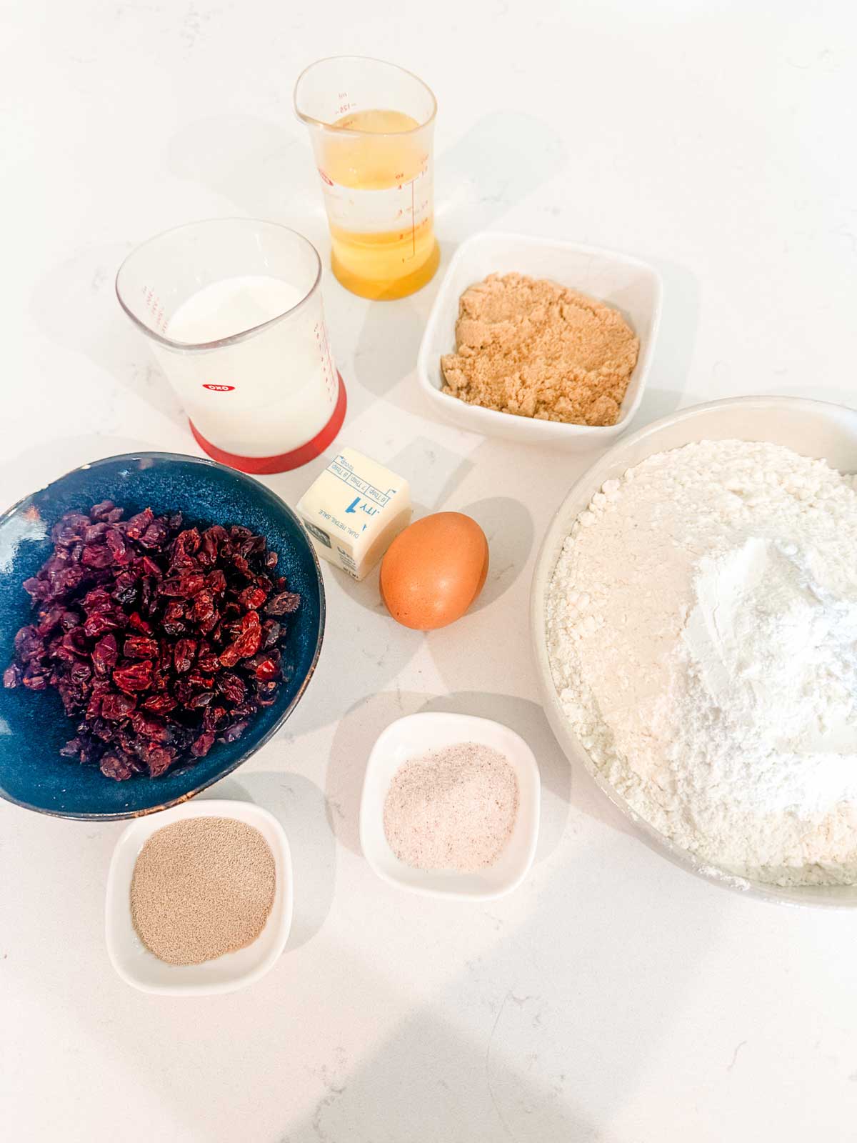 An overhead view of baking ingredients on a white surface, including flour, brown sugar, dried cranberries, an egg, a stick of butter, milk, yeast, salt, and a small cup of orange juice.