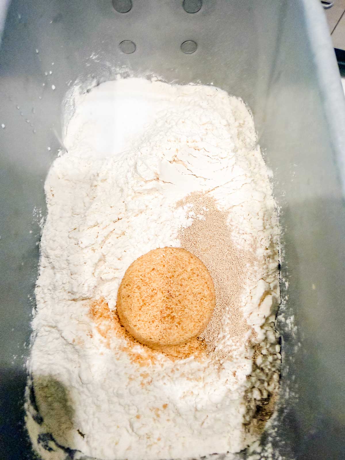 A metal bread machine pan filled with flour, a mound of brown sugar, dry yeast, and salt, ready for mixing.