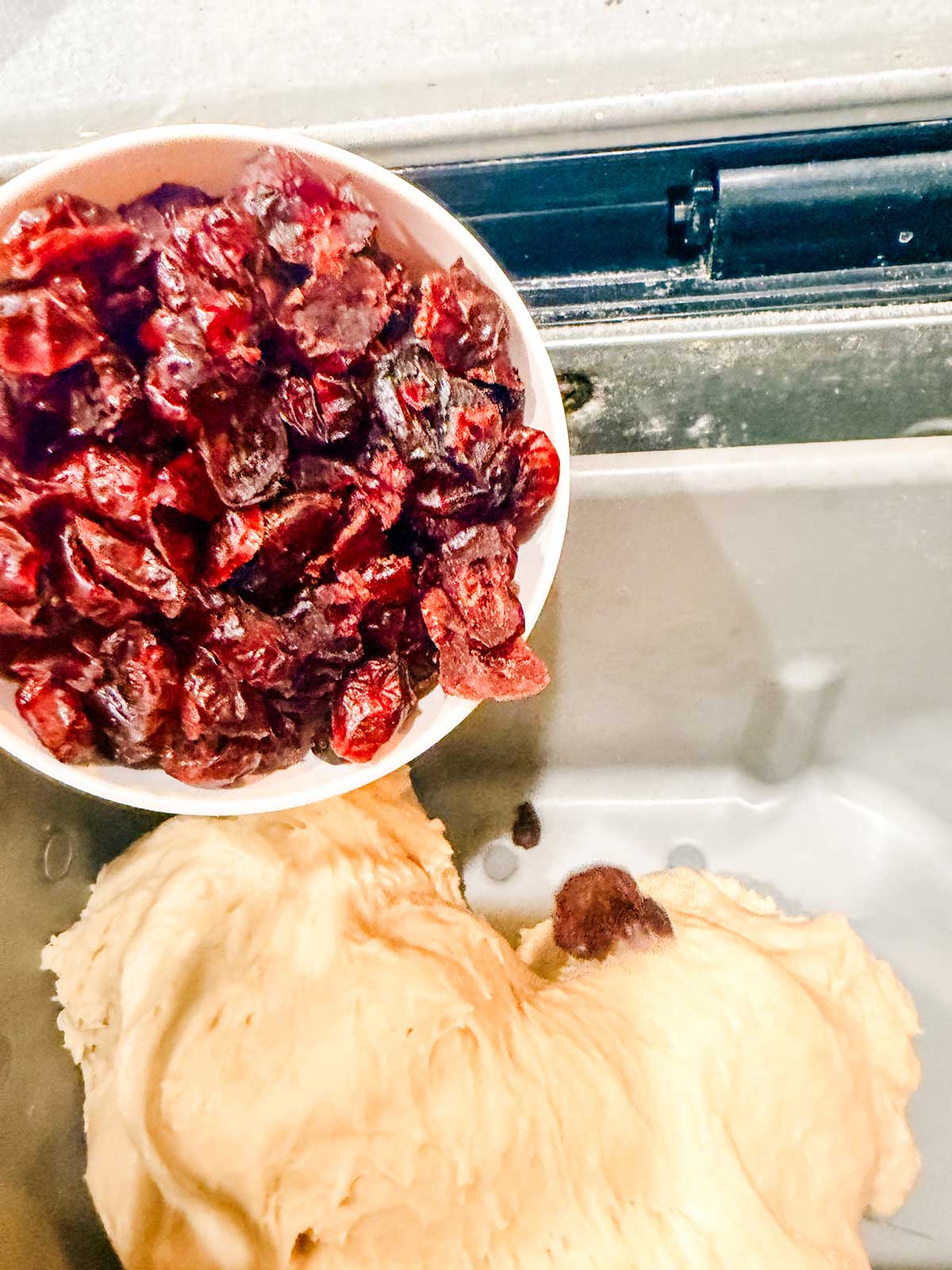 A bowl of dried cranberries being poured into a bread machine pan with kneaded dough inside.