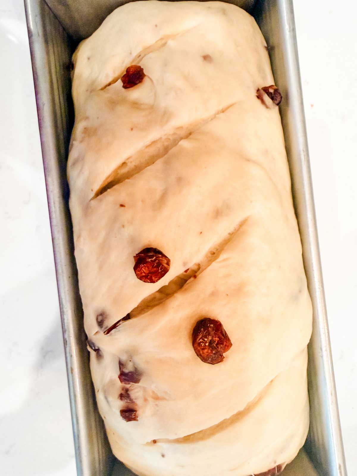 A close-up of unbaked bread dough with visible raisins, placed in a rectangular metal loaf pan and scored with three diagonal slashes on top.