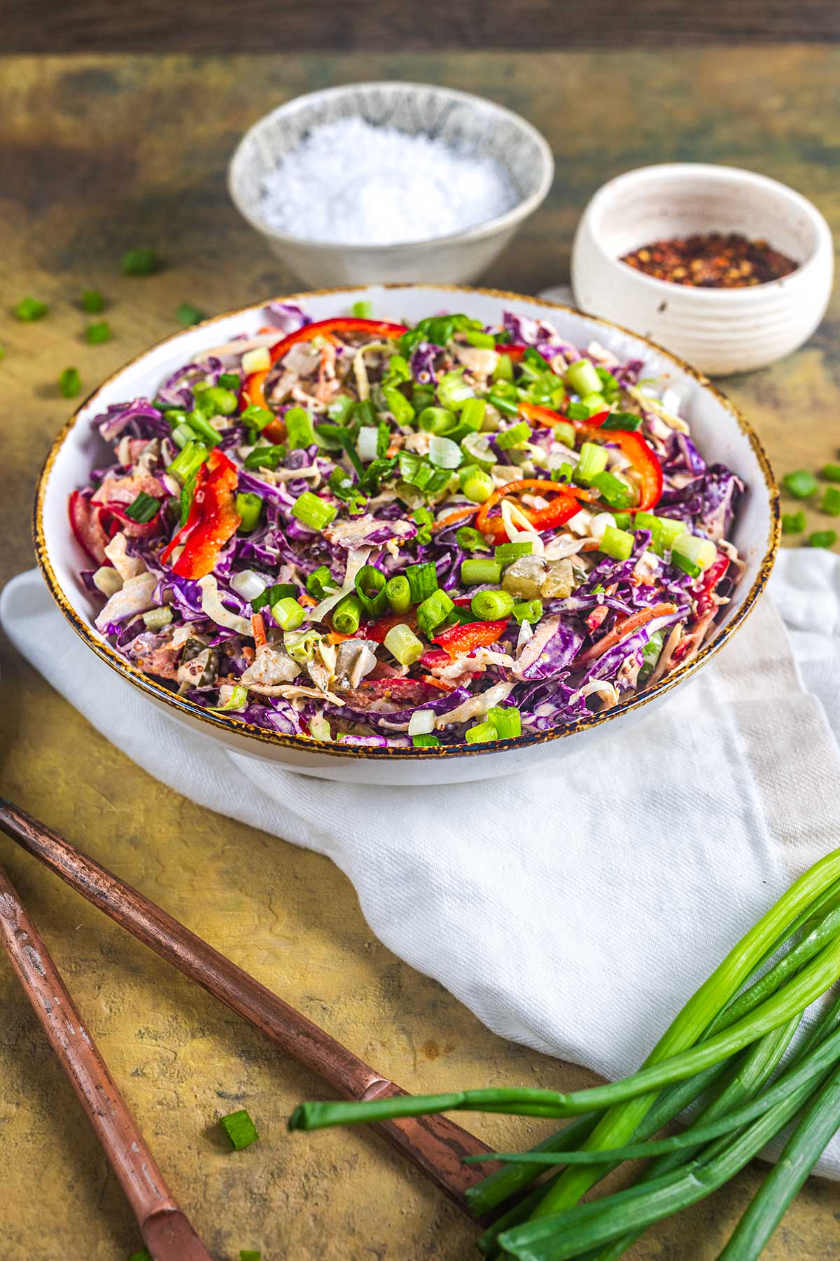 A bowl of colorful cabbage slaw with red bell peppers, green onions, and sliced almonds sits on a white napkin. Chopped green onions and wooden chopsticks are nearby, with small bowls of salt and red pepper flakes in the background.