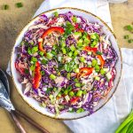 A bowl of colorful coleslaw with shredded red cabbage, sliced red bell peppers, green onions, and a creamy dressing, placed on a beige surface with utensils and a white napkin nearby.