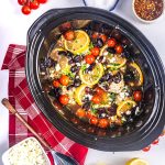A slow cooker filled with chicken breasts, lemon slices, cherry tomatoes, black olives, and herbs, surrounded by a red napkin, a bowl of feta cheese, and seasonings on a white surface.