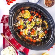 A slow cooker filled with chicken breasts, lemon slices, cherry tomatoes, black olives, and herbs, surrounded by a red napkin, a bowl of feta cheese, and seasonings on a white surface.