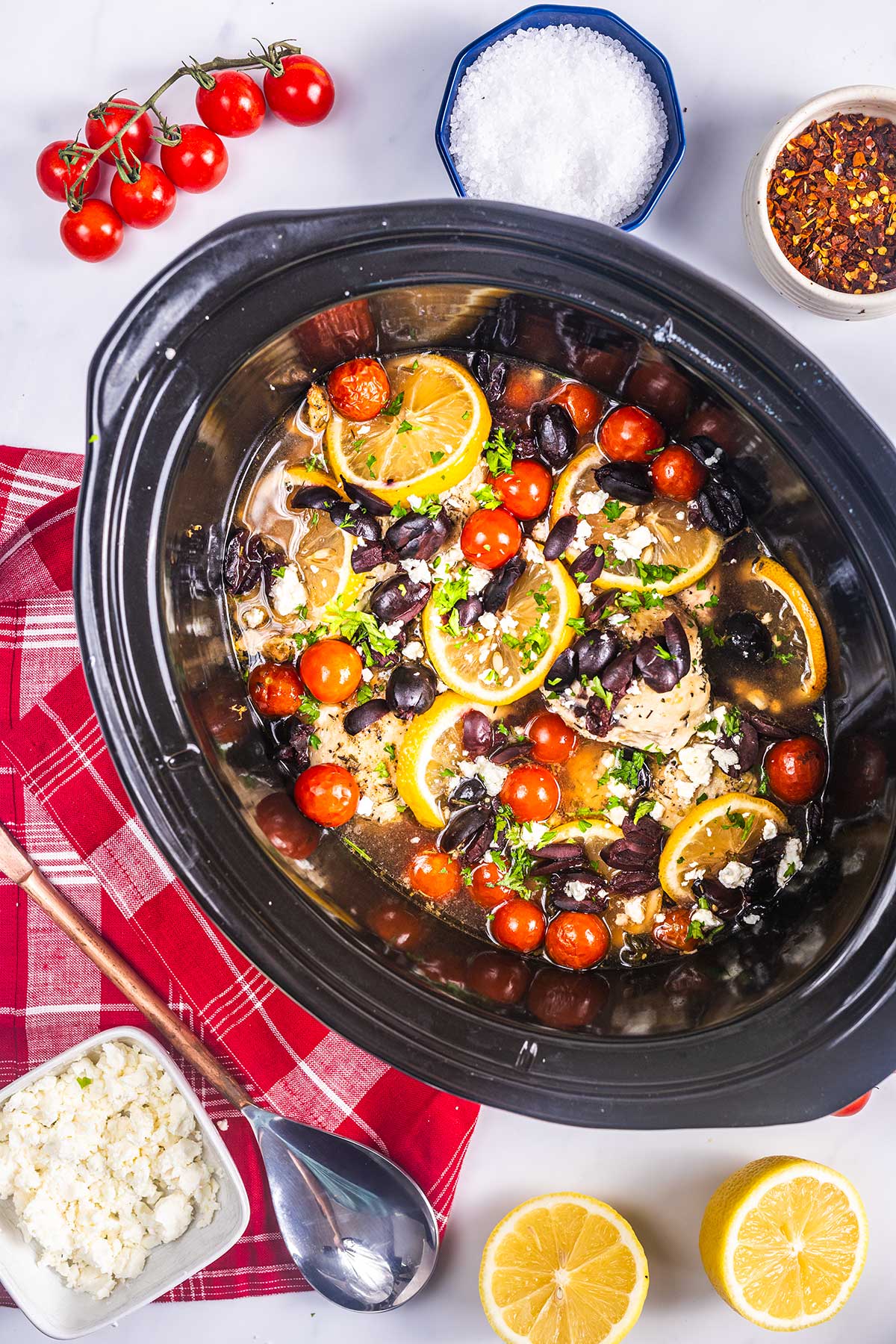 A slow cooker filled with chicken breasts topped with lemon slices, cherry tomatoes, olives, and herbs. Surrounding the slow cooker are bowls of feta cheese, salt, red pepper flakes, a spoon, lemons, and a red plaid napkin.