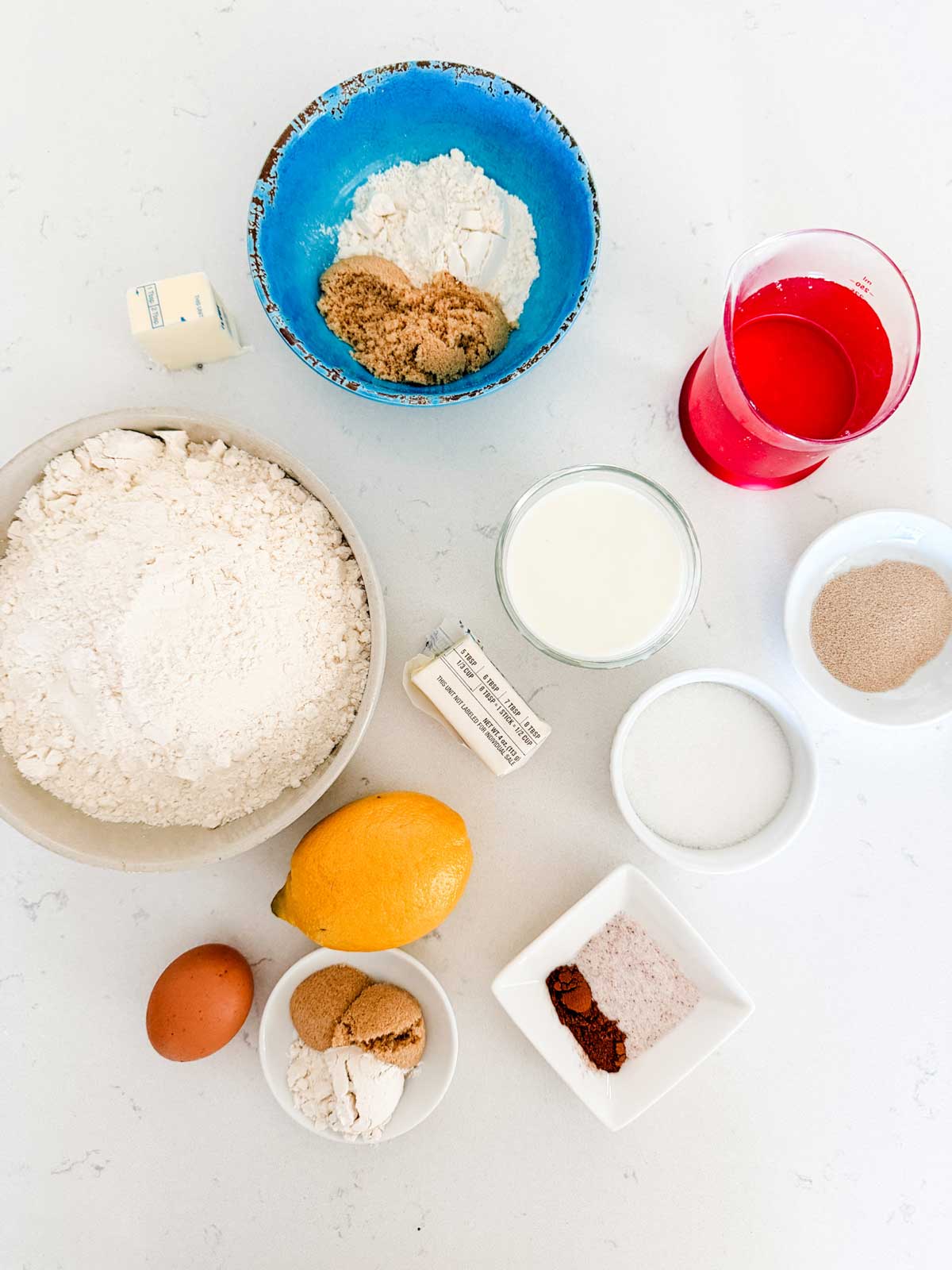 A top-down view of baking ingredients on a white surface, including flour, brown sugar, butter, milk, red liquid, yeast, lemon, an egg, and small bowls with spices, salt, and sugar.