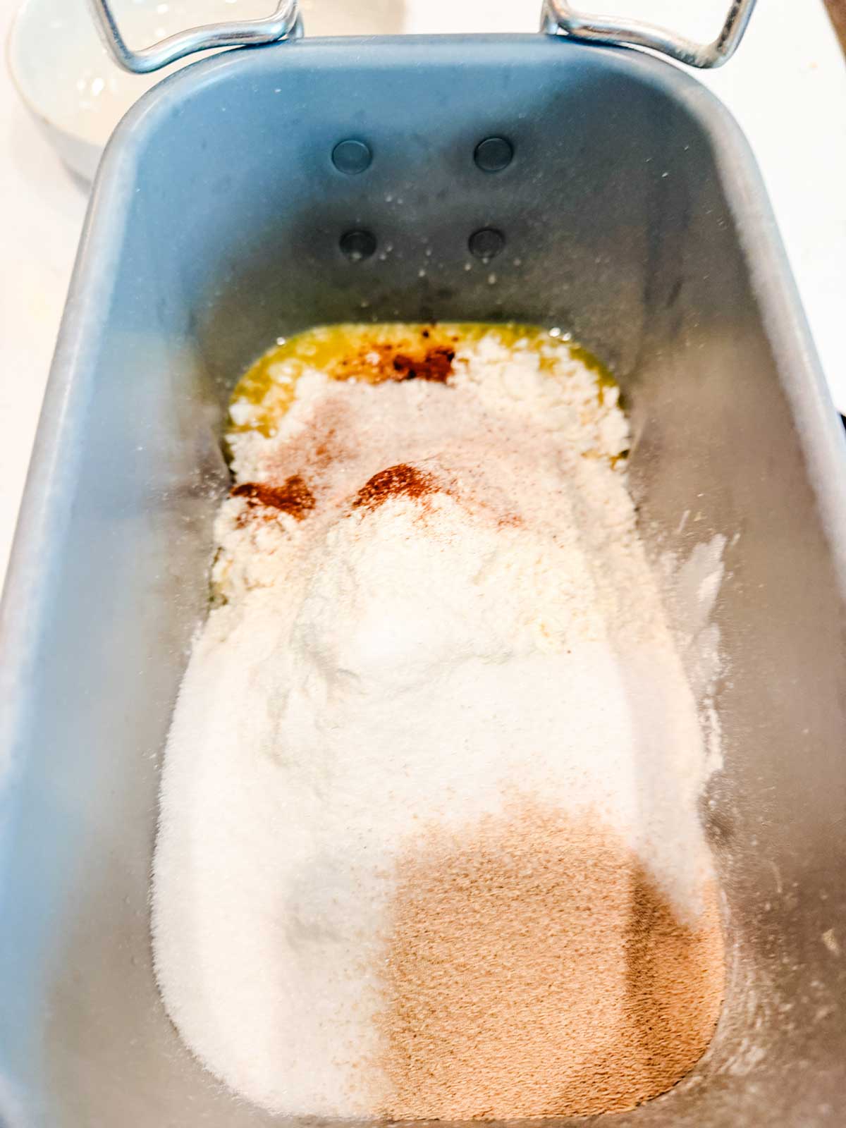 A bread machine pan filled with flour, sugar, yeast, and other dry ingredients, ready to be mixed for bread making.