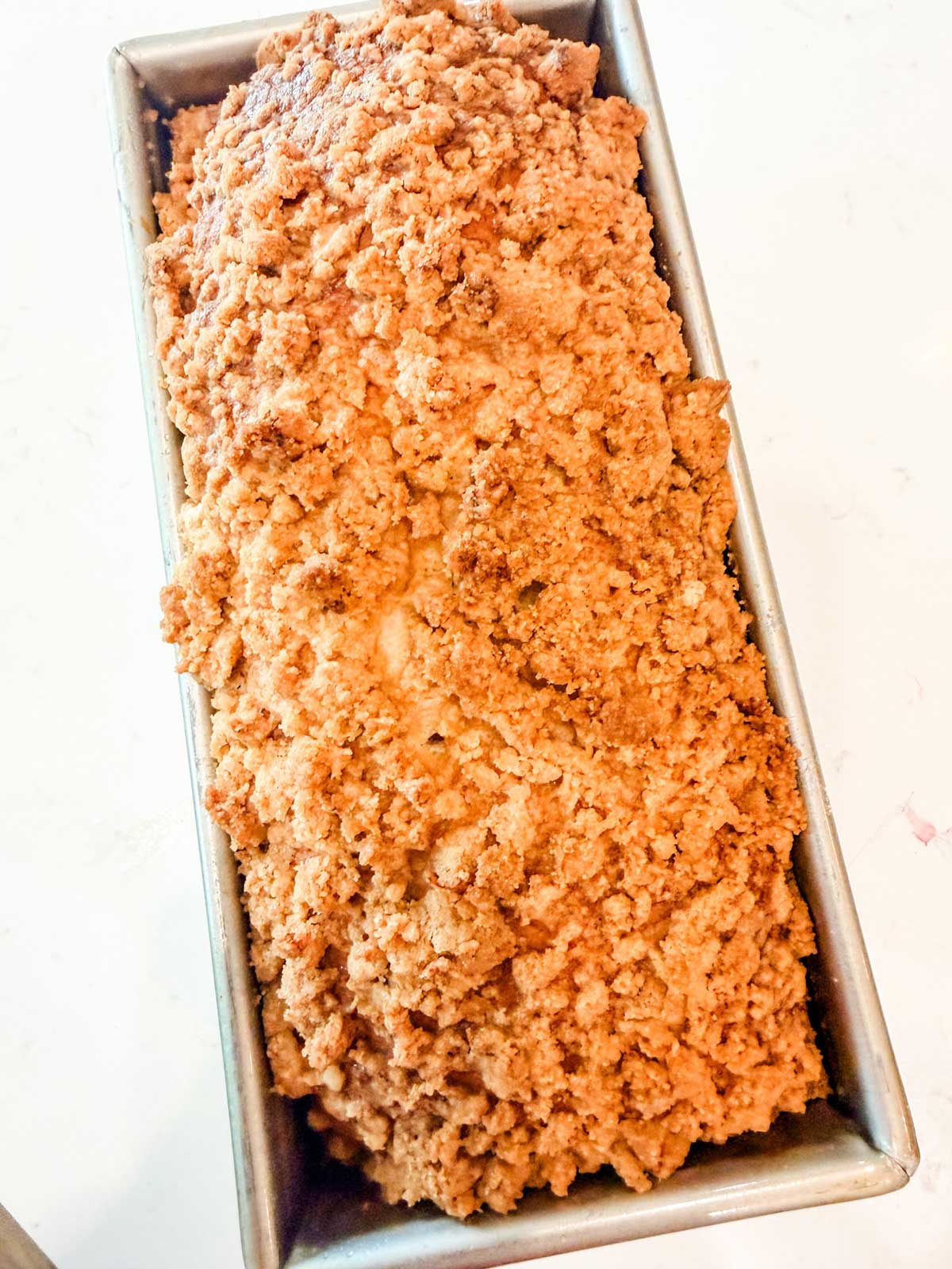 A close-up of a golden-brown loaf with a crumbly, textured top, baked in a rectangular metal loaf pan on a light-colored surface.