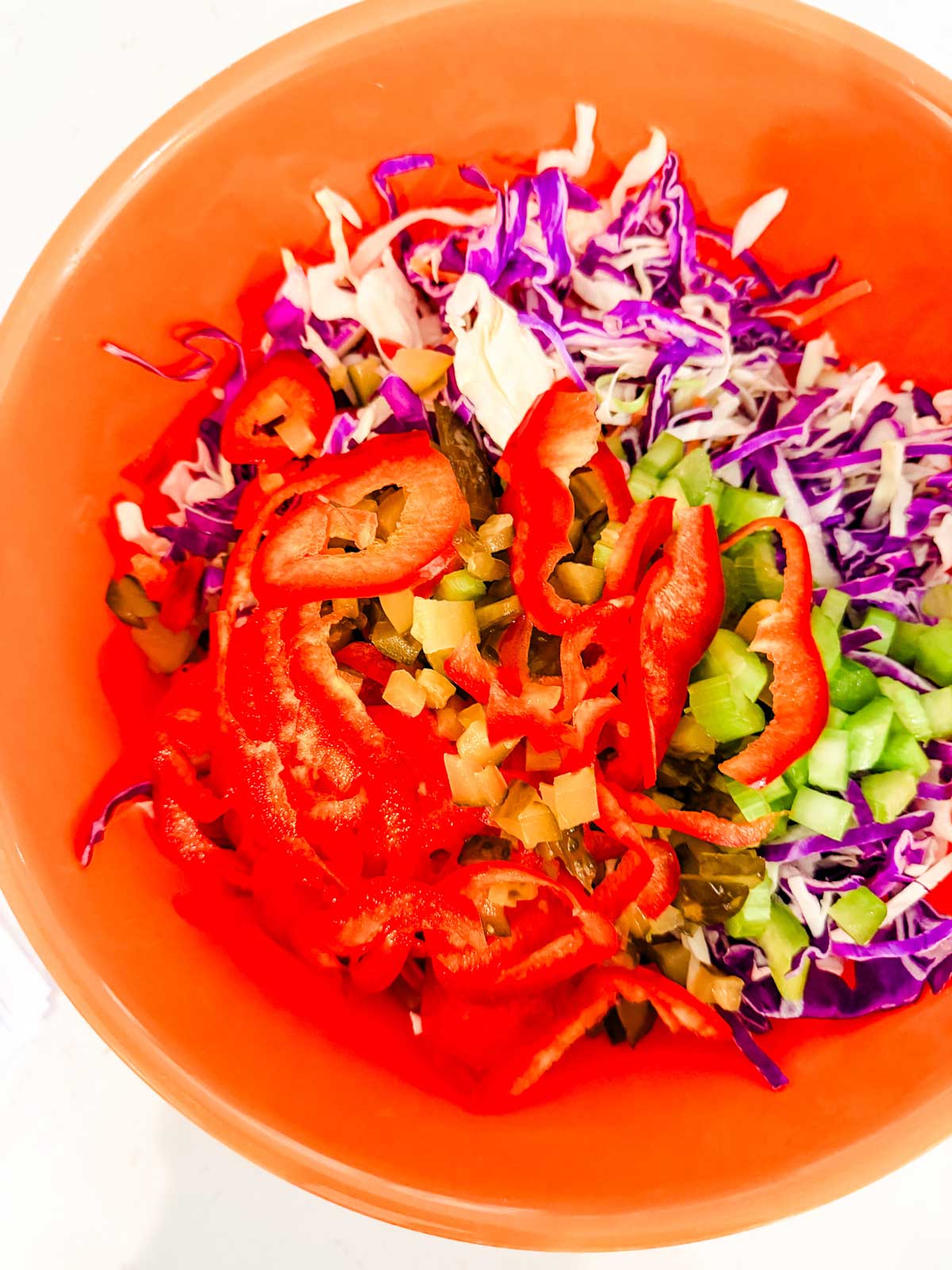 A bright orange bowl filled with shredded purple cabbage, sliced red bell peppers, green celery, and diced pickles.