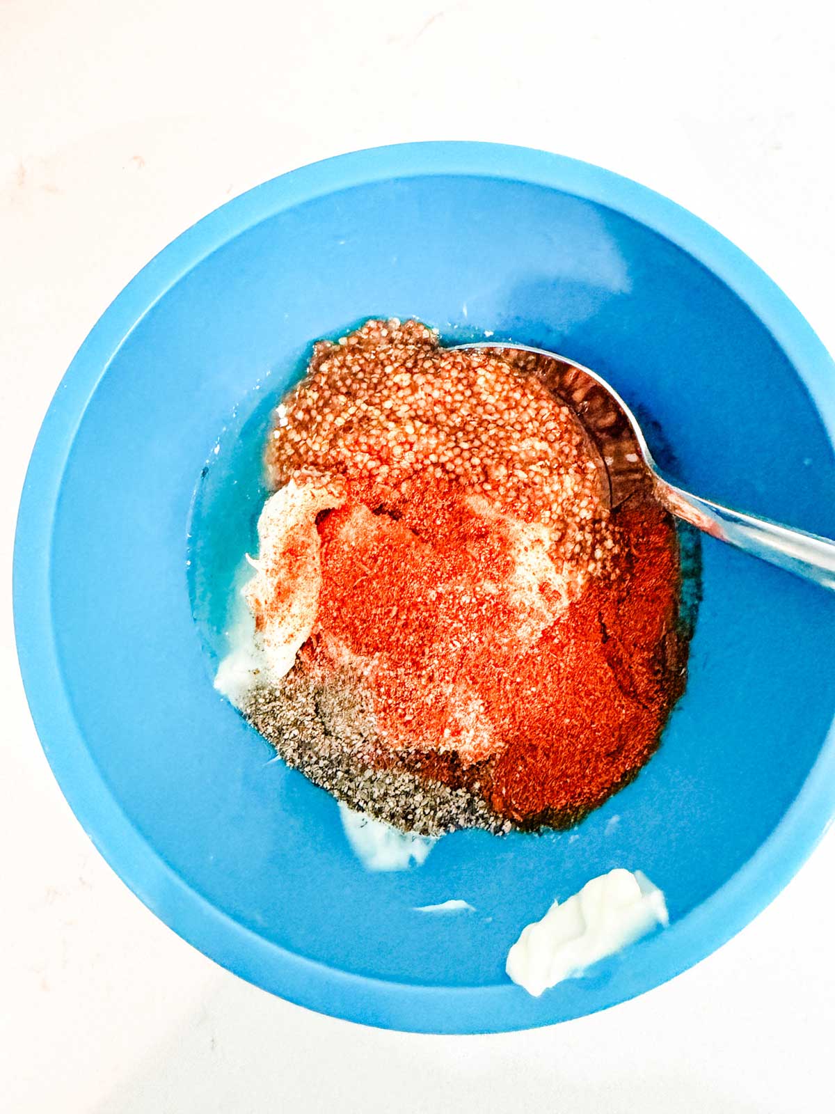 A blue bowl containing a mixture of spices, mustard, ground black pepper, paprika, and white creamy ingredients, with a spoon resting inside.