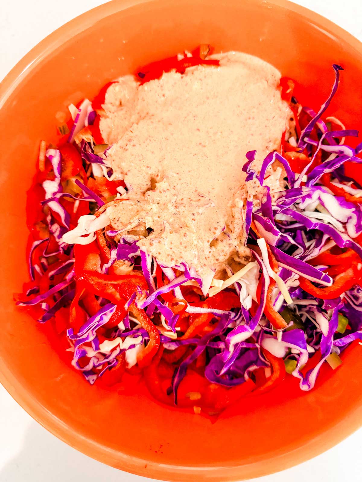 A bright orange bowl filled with shredded red cabbage, sliced red bell peppers, and a creamy, light-colored dressing on top.
