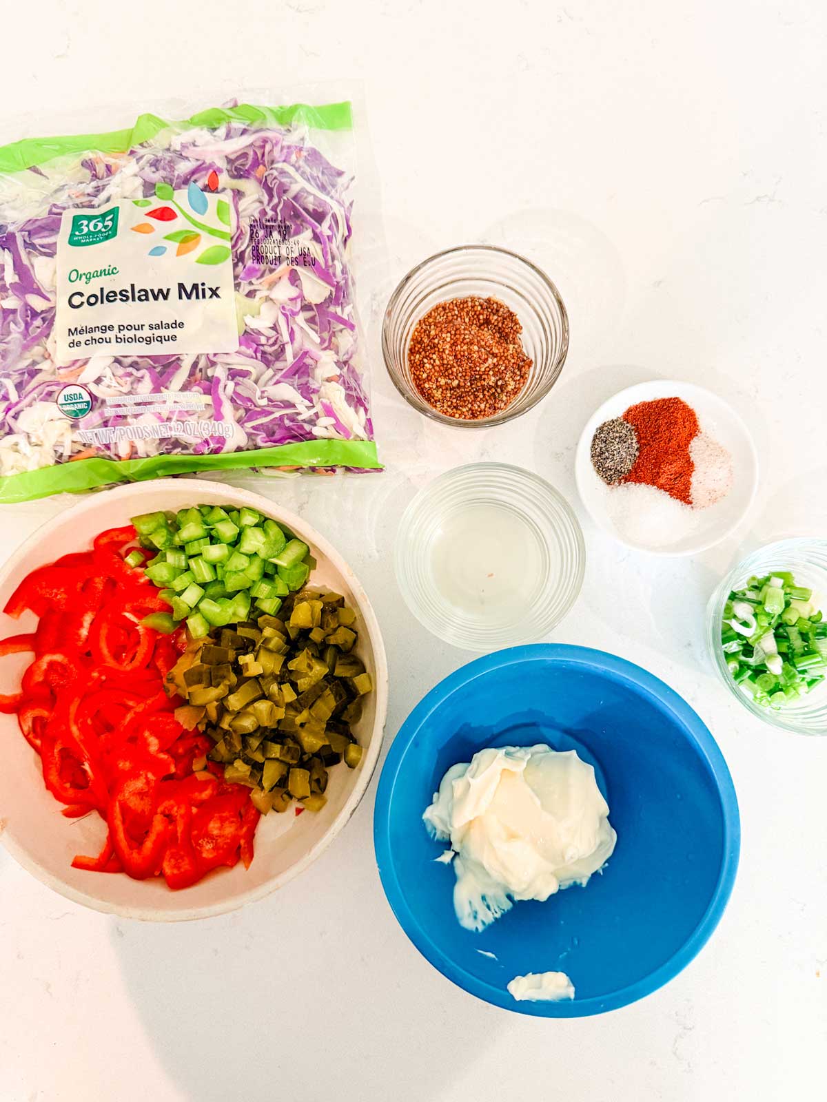 A variety of ingredients on a white surface: a bag of coleslaw mix, bowls with spices, sliced celery, pickles, tomatoes, mayonnaise, green onions, and a small bowl of clear liquid.