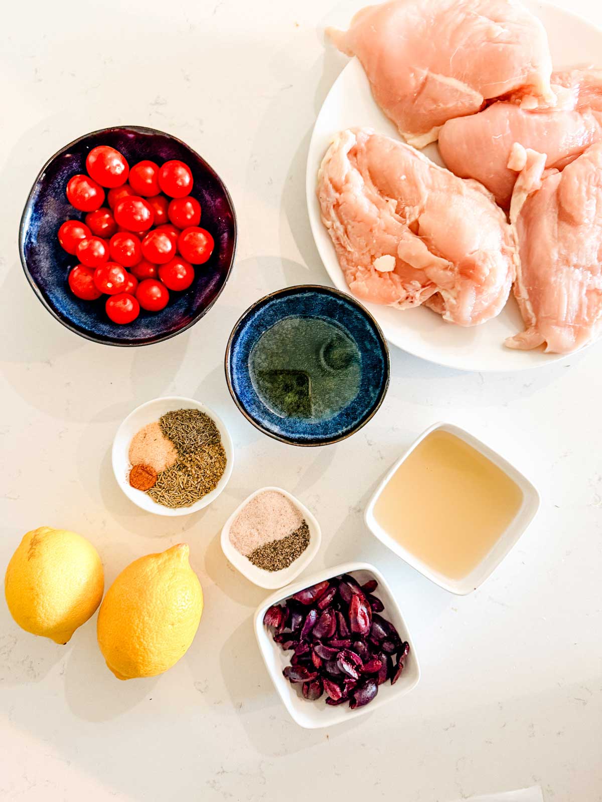 A top-down view of raw chicken breasts, cherry tomatoes, kalamata olives, two lemons, small bowls of spices, salt, pepper, olive oil, and a square dish of broth on a white surface.