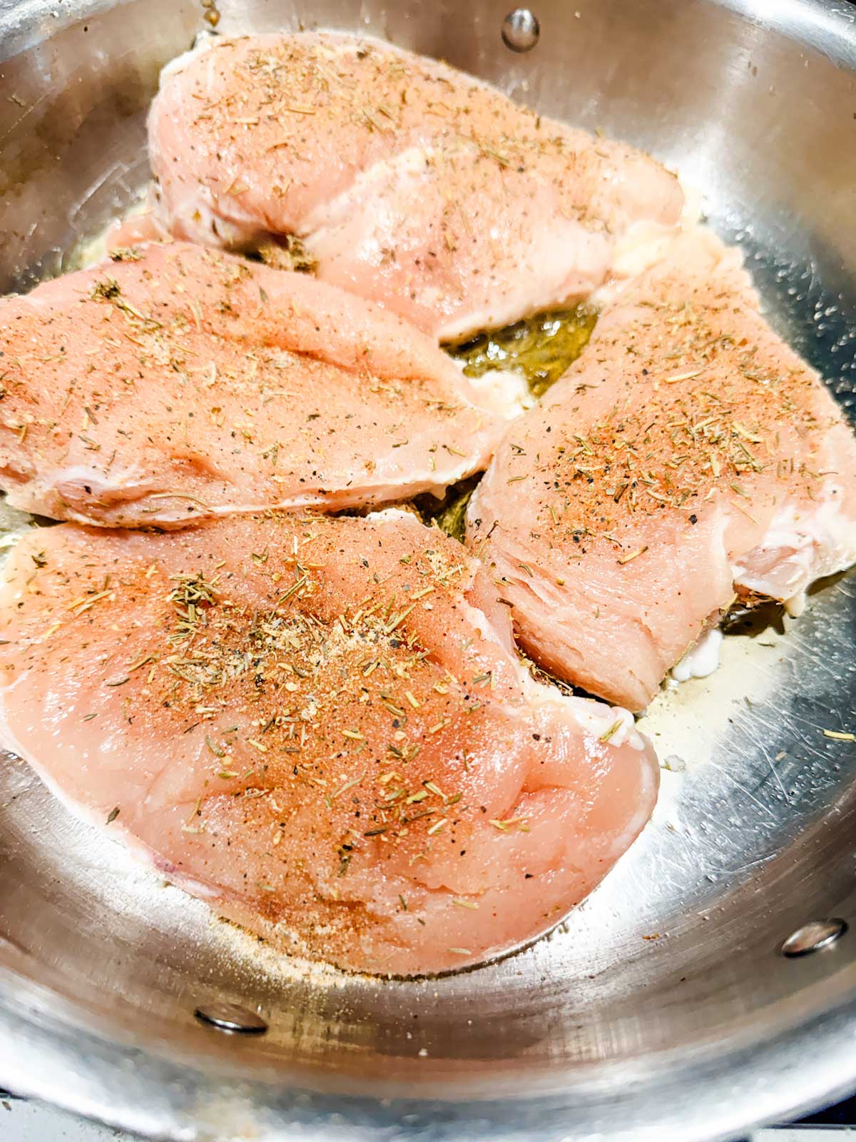Raw, seasoned chicken breasts are placed in a stainless steel pan with a bit of oil, ready to be cooked. The chicken is sprinkled with various spices and herbs.