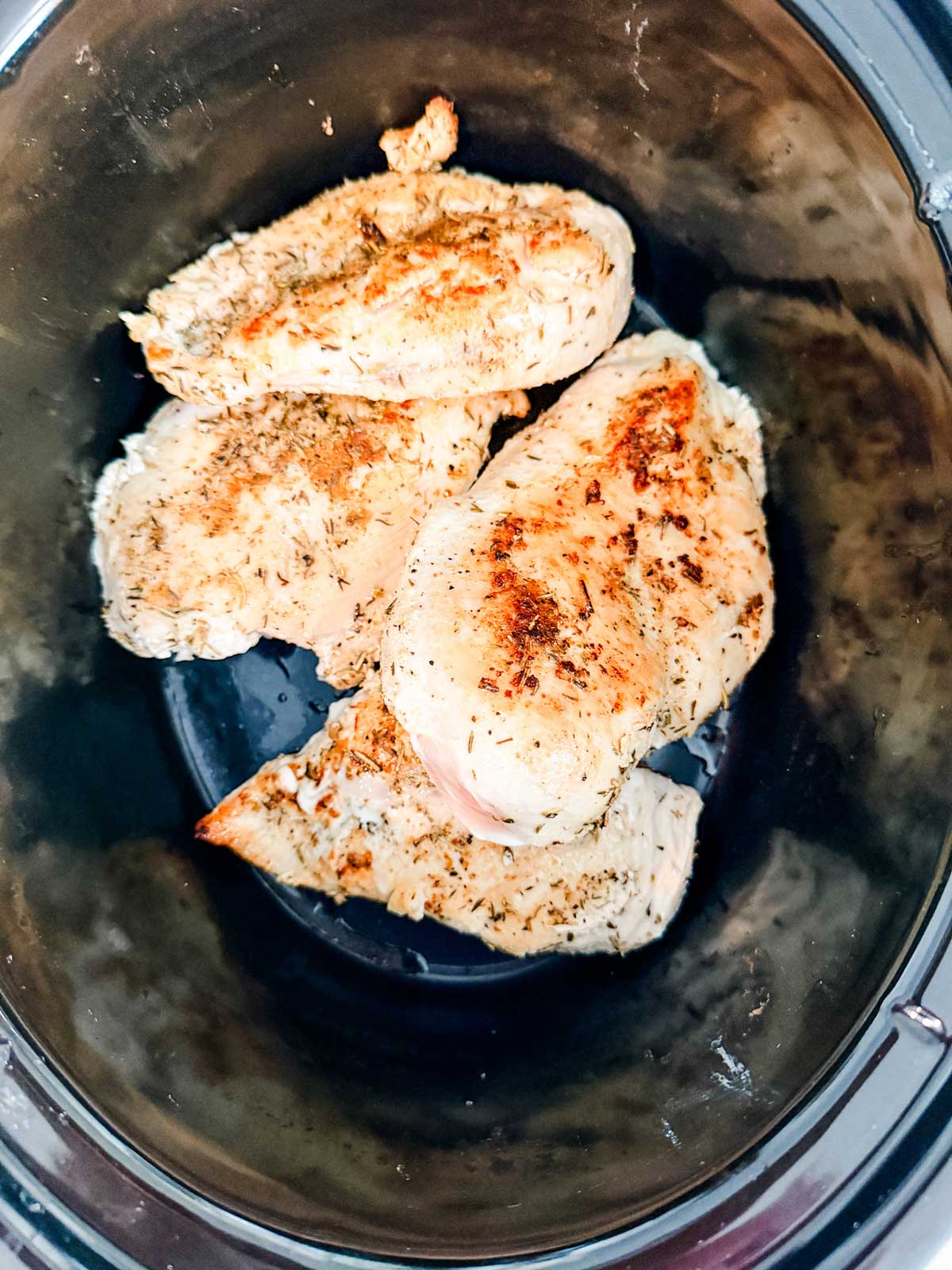 Four seasoned, cooked chicken breasts rest in a dark slow cooker, showing light browning and visible herbs on the surface.