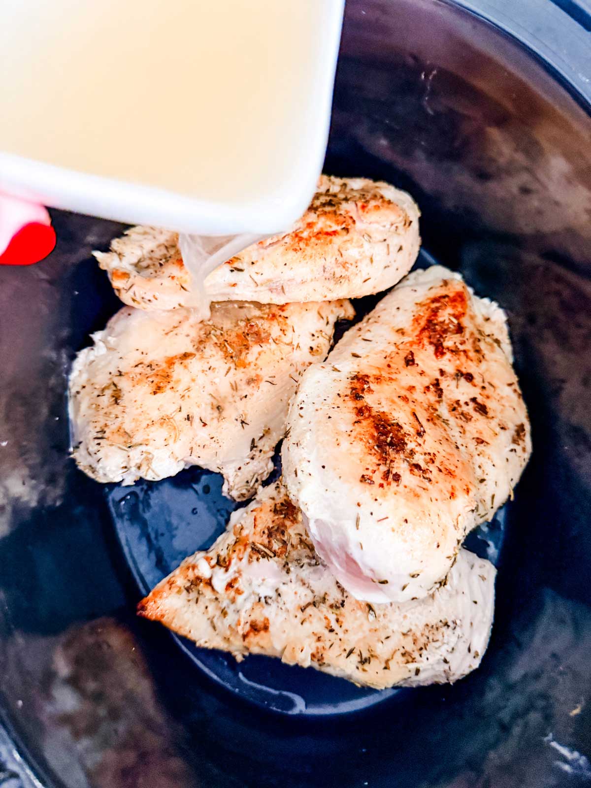 Seasoned chicken breasts in a slow cooker with broth being poured over them from a white container. The chicken appears browned and ready for slow cooking.