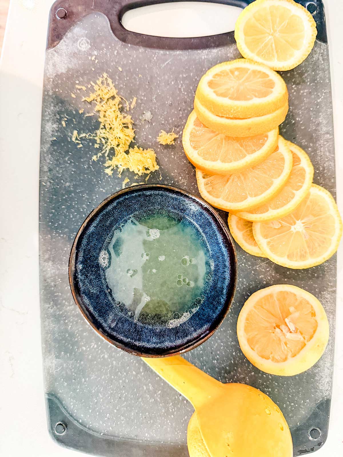 A cutting board with lemon slices, lemon zest, a halved lemon, and a small bowl of lemon juice with a yellow juicer.