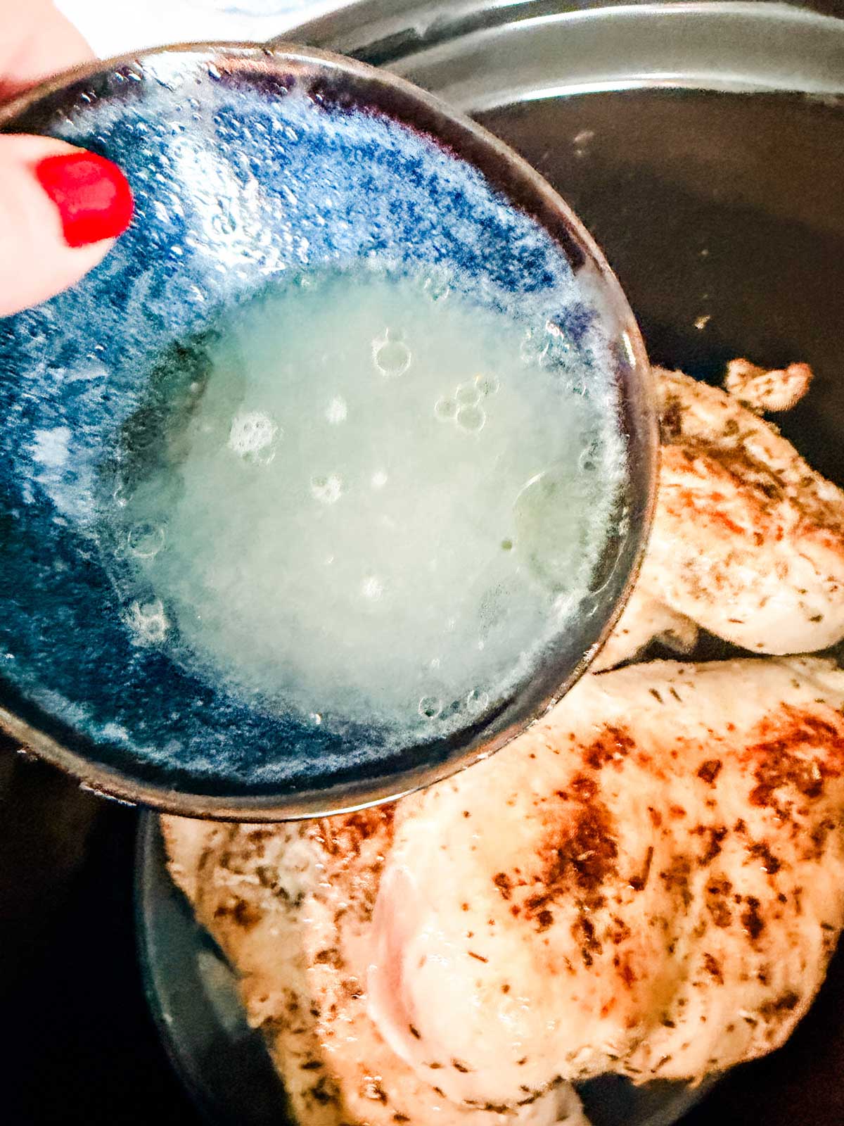 A hand with red nail polish holds a blue bowl of liquid over cooked, seasoned chicken pieces in a pan, preparing to pour the liquid onto the chicken.