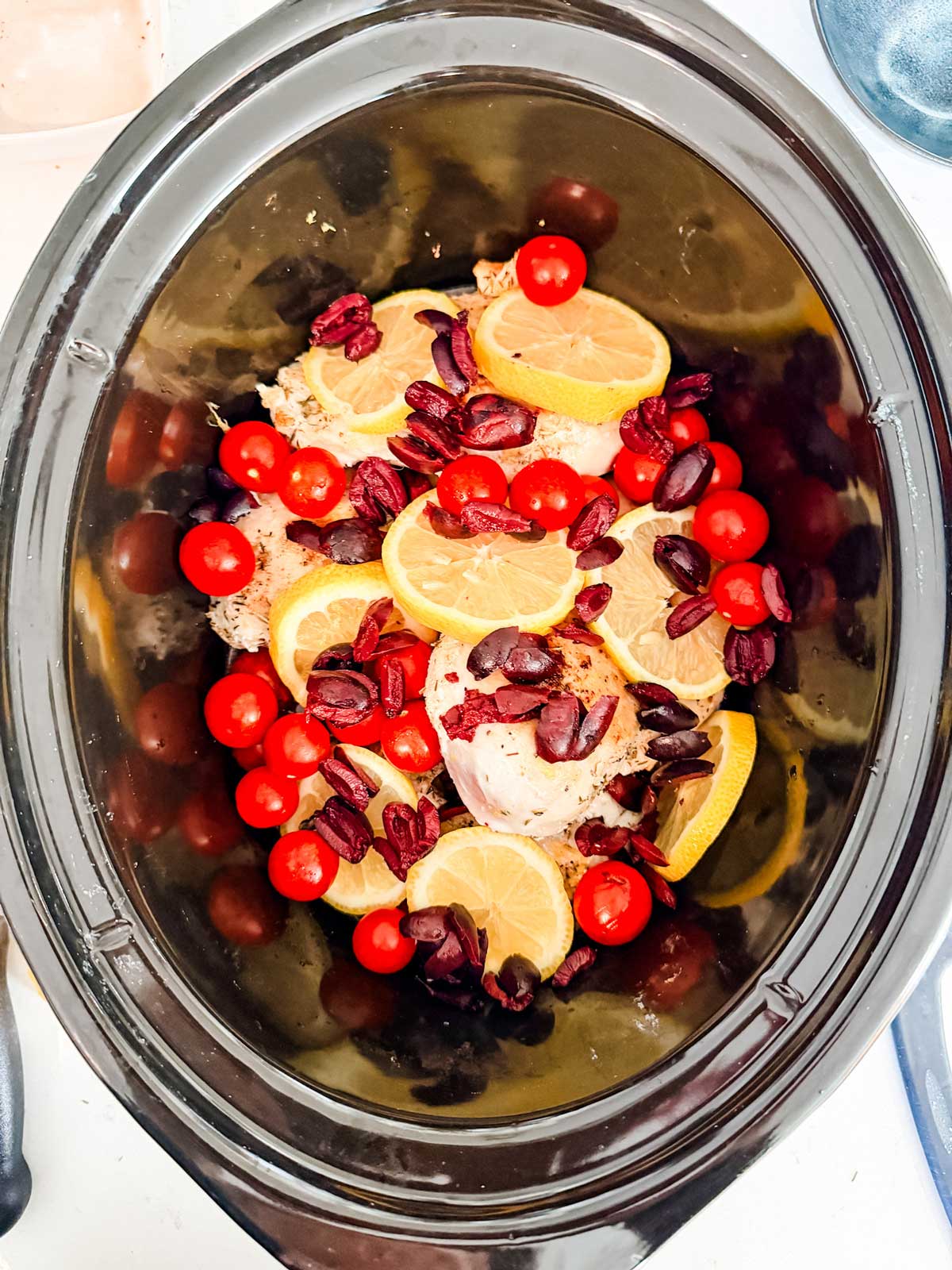 Chicken breasts in a slow cooker topped with lemon slices, cherry tomatoes, and chopped kalamata olives, ready to be cooked.