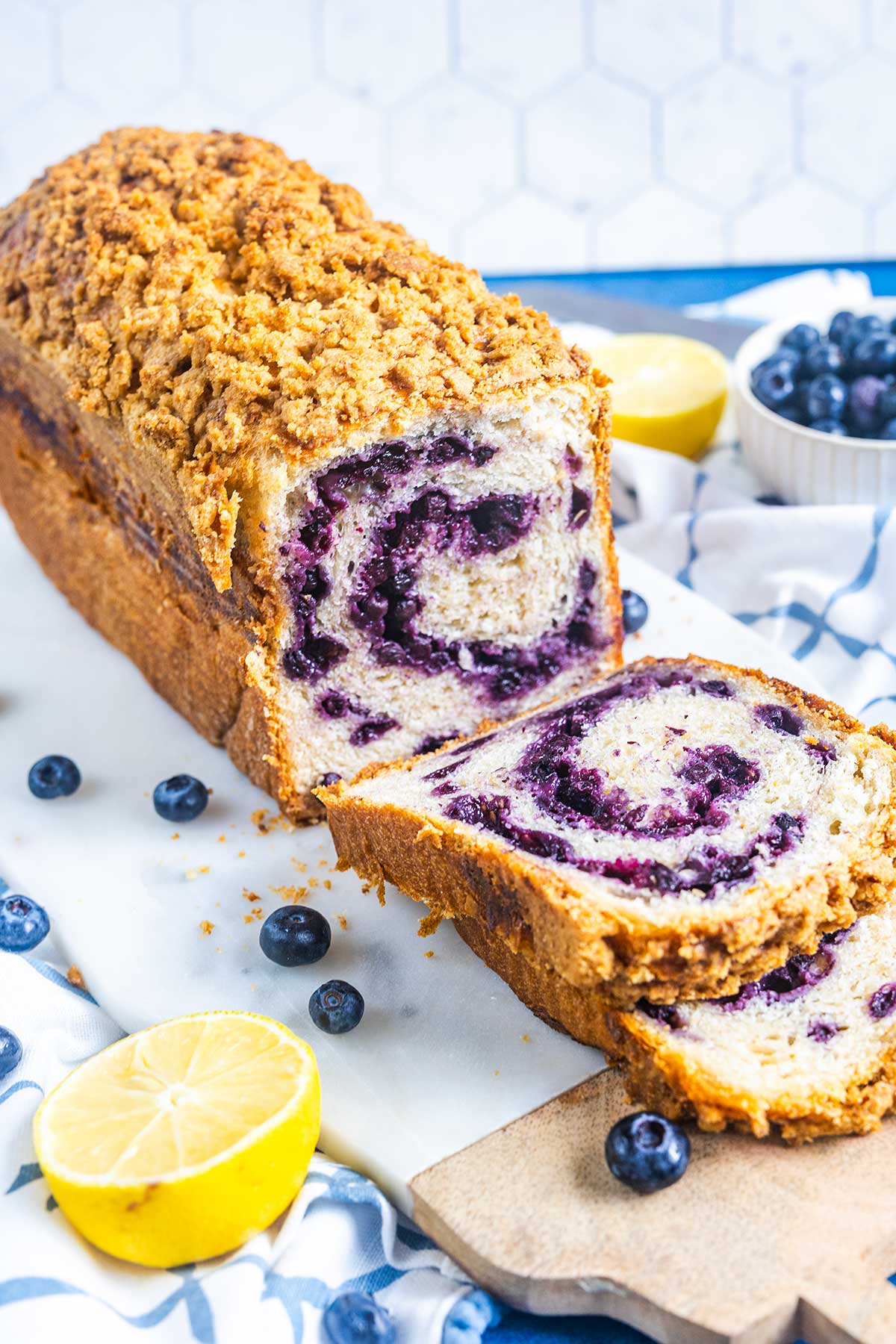 A sliced loaf of blueberry swirl bread topped with crumb streusel sits on a wooden board. Fresh blueberries and lemon halves are scattered nearby on a white cloth.