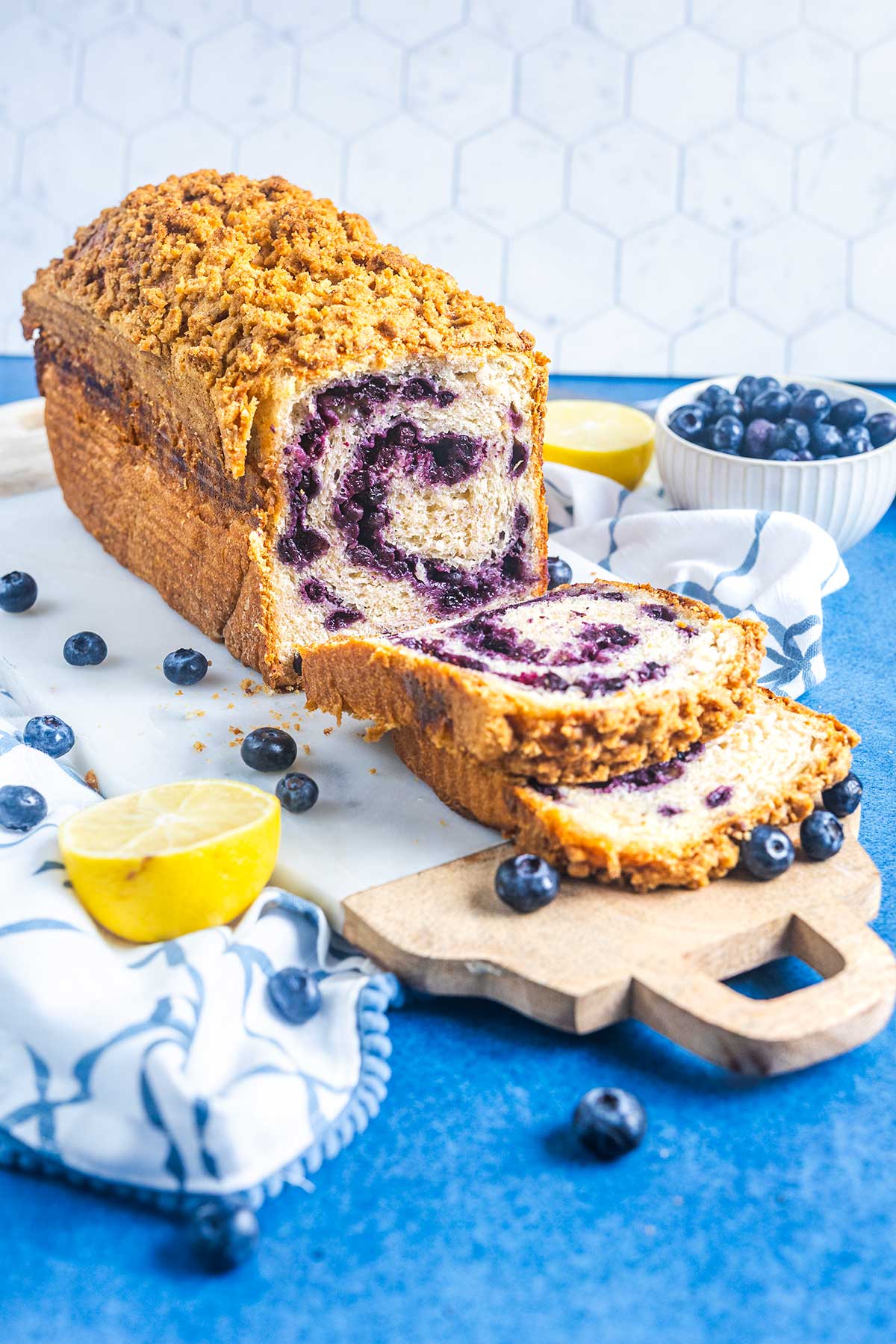 A sliced loaf of blueberry swirl bread sits on a wooden board, surrounded by fresh blueberries and a halved lemon on a blue surface, with a bowl of blueberries and a white cloth nearby.