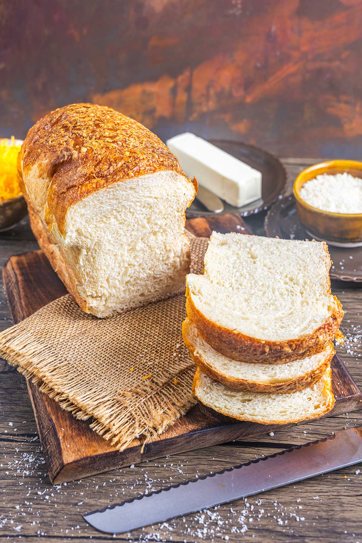 A loaf of homemade bread sits on a wooden cutting board, partially sliced, with three bread slices in front. Nearby are a serrated knife, a stick of butter, and a small bowl of flour on a rustic wooden table.