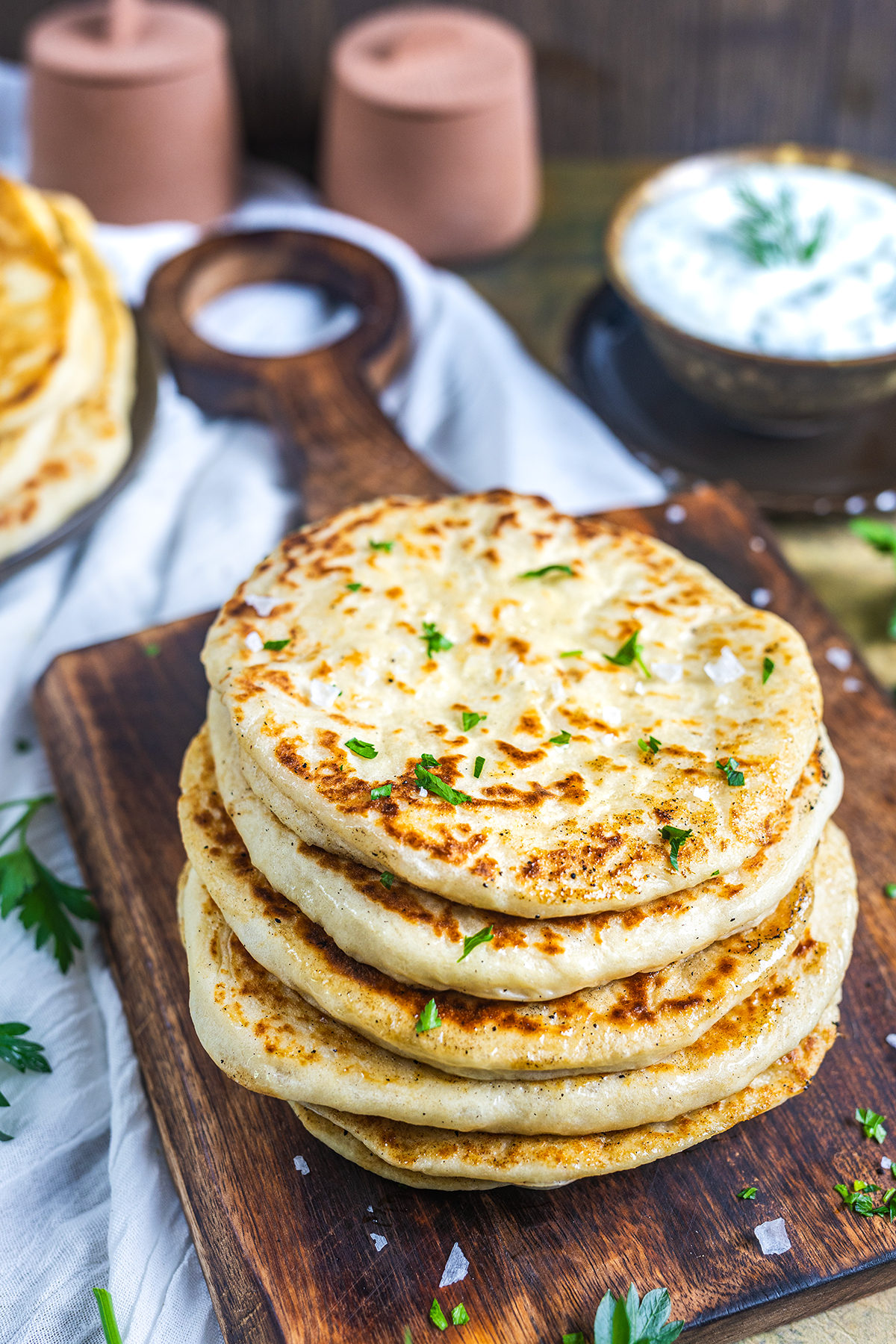 A stack of golden, pan-fried flatbreads garnished with fresh herbs sits on a wooden serving board. In the background, a bowl of creamy dip with herbs and a plate of more flatbreads are visible.