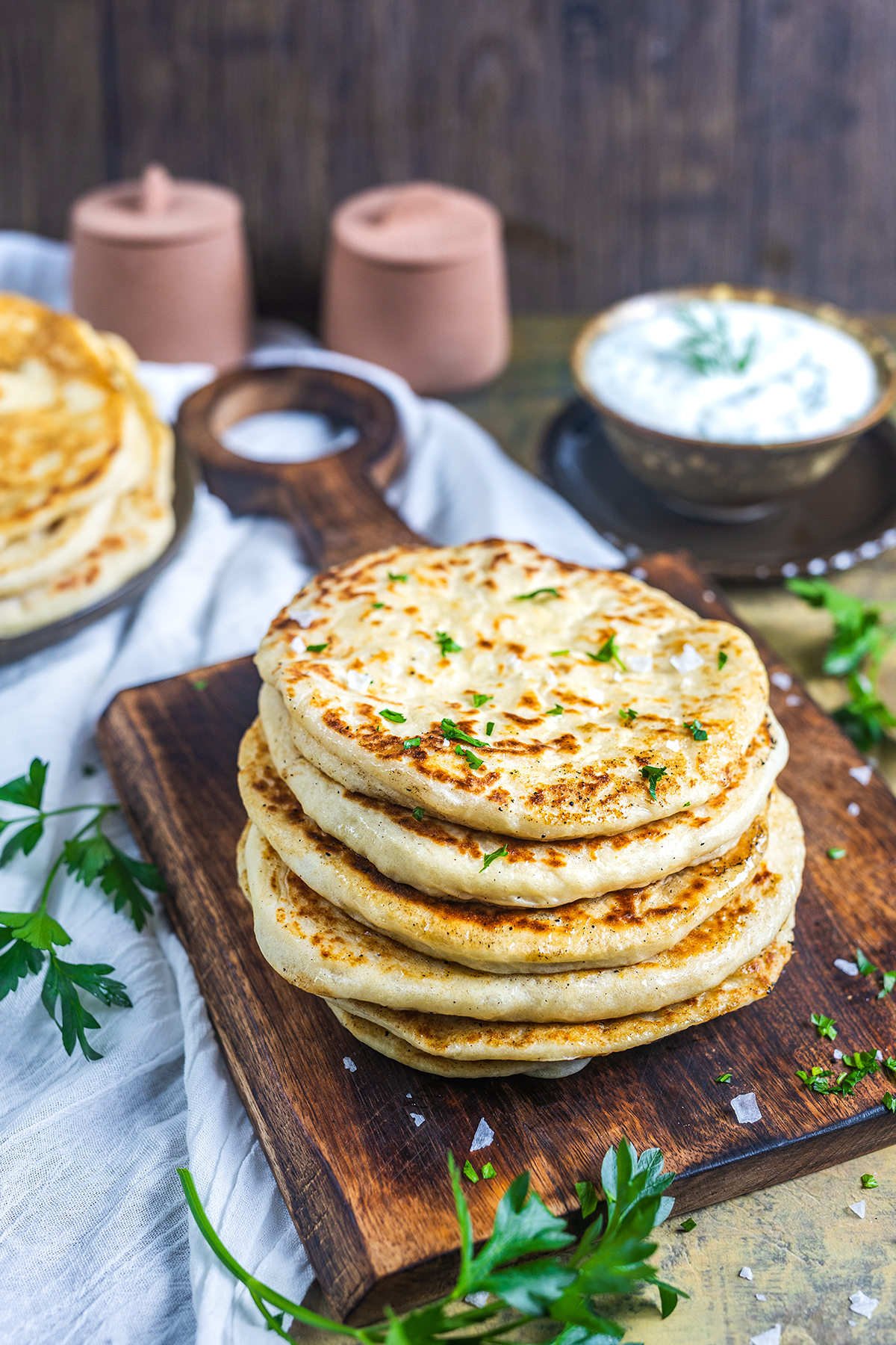 A stack of golden, fluffy flatbreads garnished with chopped herbs sits on a wooden board, surrounded by fresh parsley, with a bowl of white sauce and more flatbreads in the background.