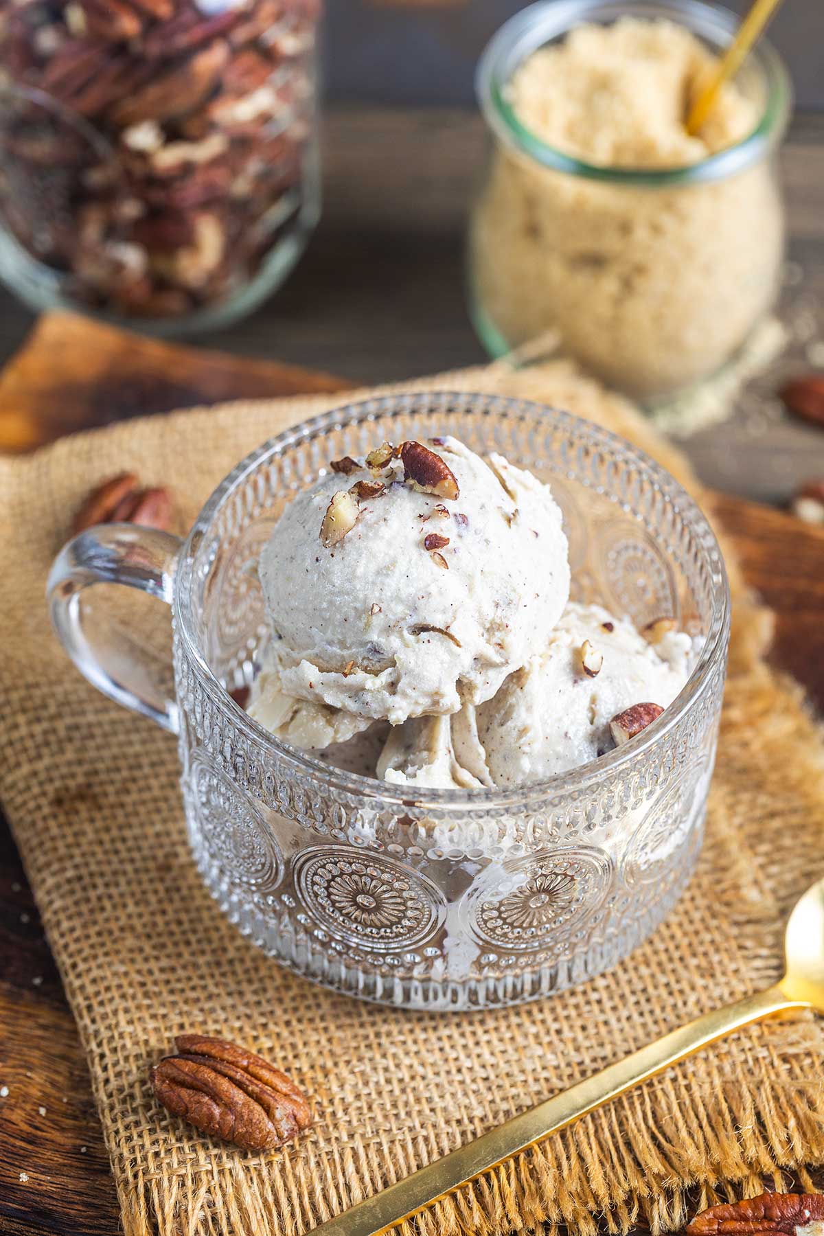 A glass cup filled with two scoops of pecan ice cream sits on a burlap cloth, surrounded by pecans. In the background are jars of brown sugar and pecans, with a gold spoon nearby.