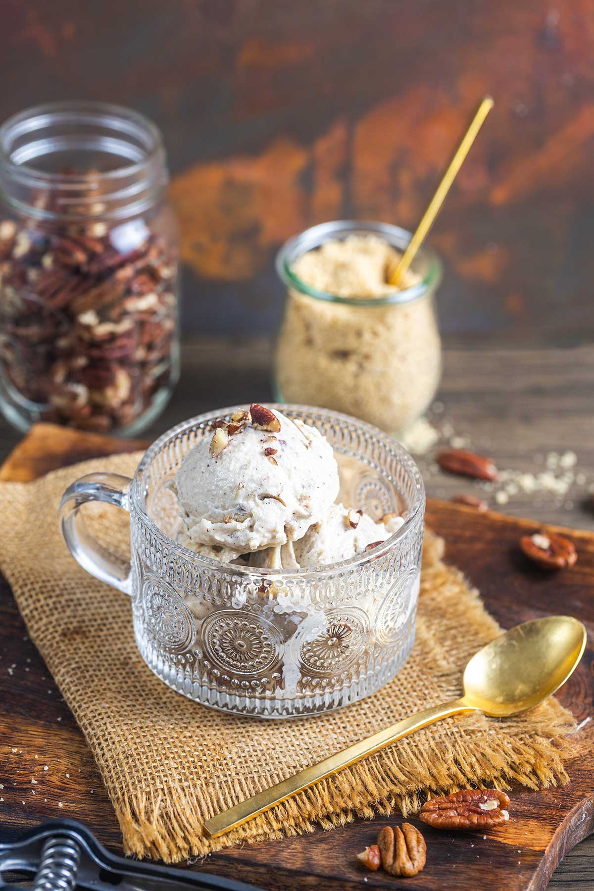 A glass cup filled with scoops of pecan ice cream sits on a burlap mat, surrounded by pecans, a gold spoon, a jar of brown sugar, and a jar of pecans on a wooden surface.