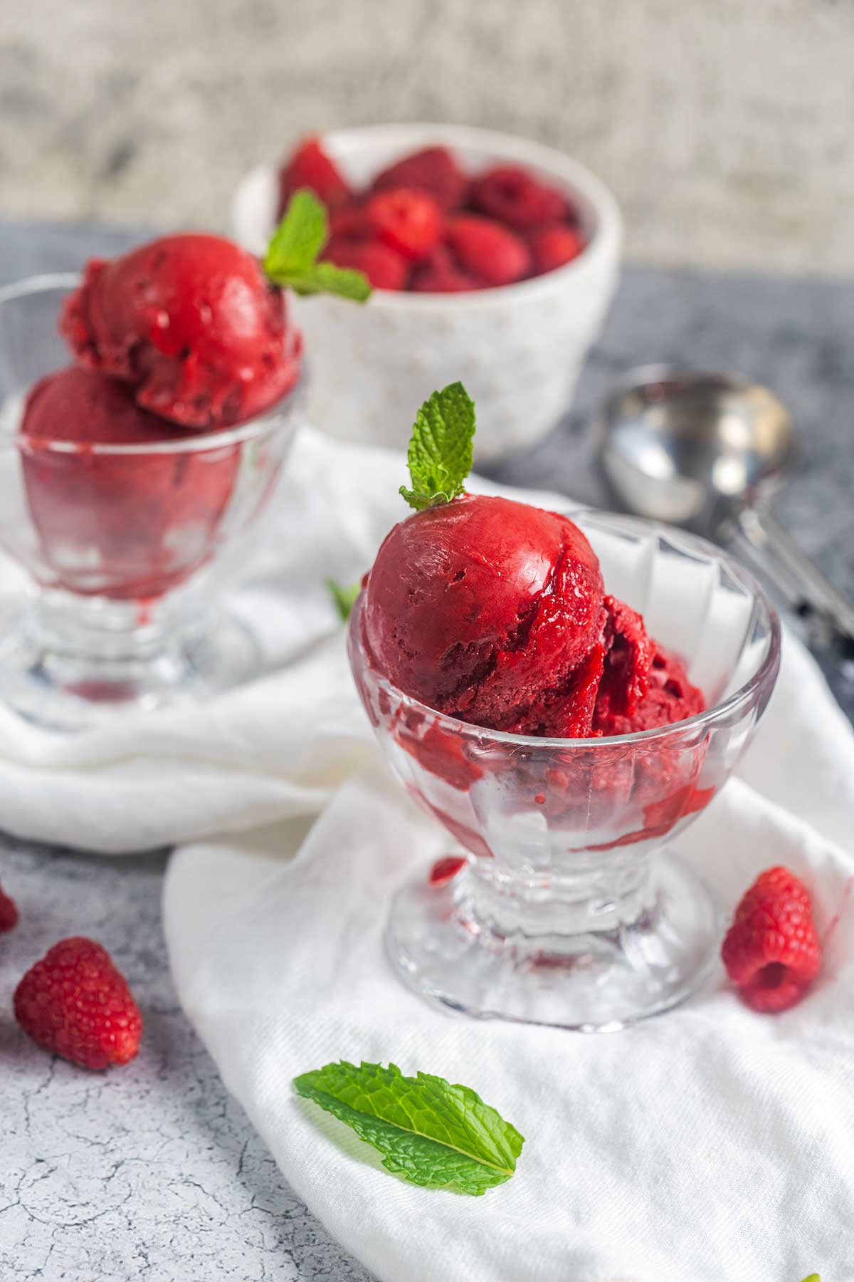 Two glass bowls filled with scoops of vibrant red raspberry sorbet, garnished with fresh mint leaves. Loose raspberries and a white bowl of berries are in the background, next to metal ice cream scoops on a white cloth.