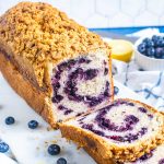 A loaf of blueberry swirl bread with a crumbly streusel topping sits on a cutting board. One slice is cut, showing the blueberry swirl inside. Fresh blueberries and a lemon are in the background.
