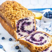 A loaf of blueberry swirl bread with a crumbly streusel topping sits on a cutting board. One slice is cut, showing the blueberry swirl inside. Fresh blueberries and a lemon are in the background.