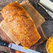 A golden-brown loaf of bread on a wooden board with a knife nearby, shredded cheese in a bowl, and a stick of butter in the background.