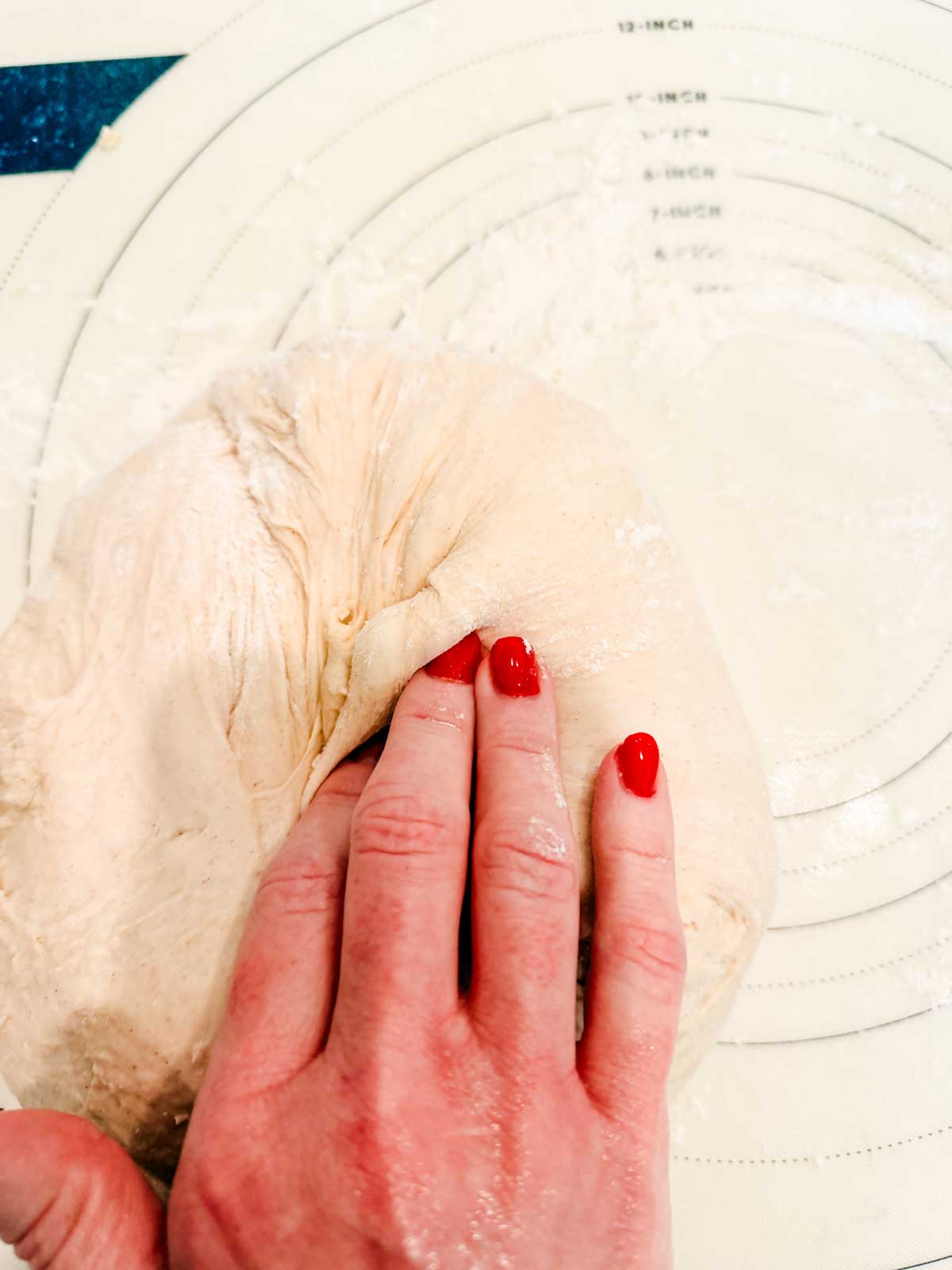 A hand with red nail polish presses into a ball of bread dough on a floured surface with circular measurement markings.