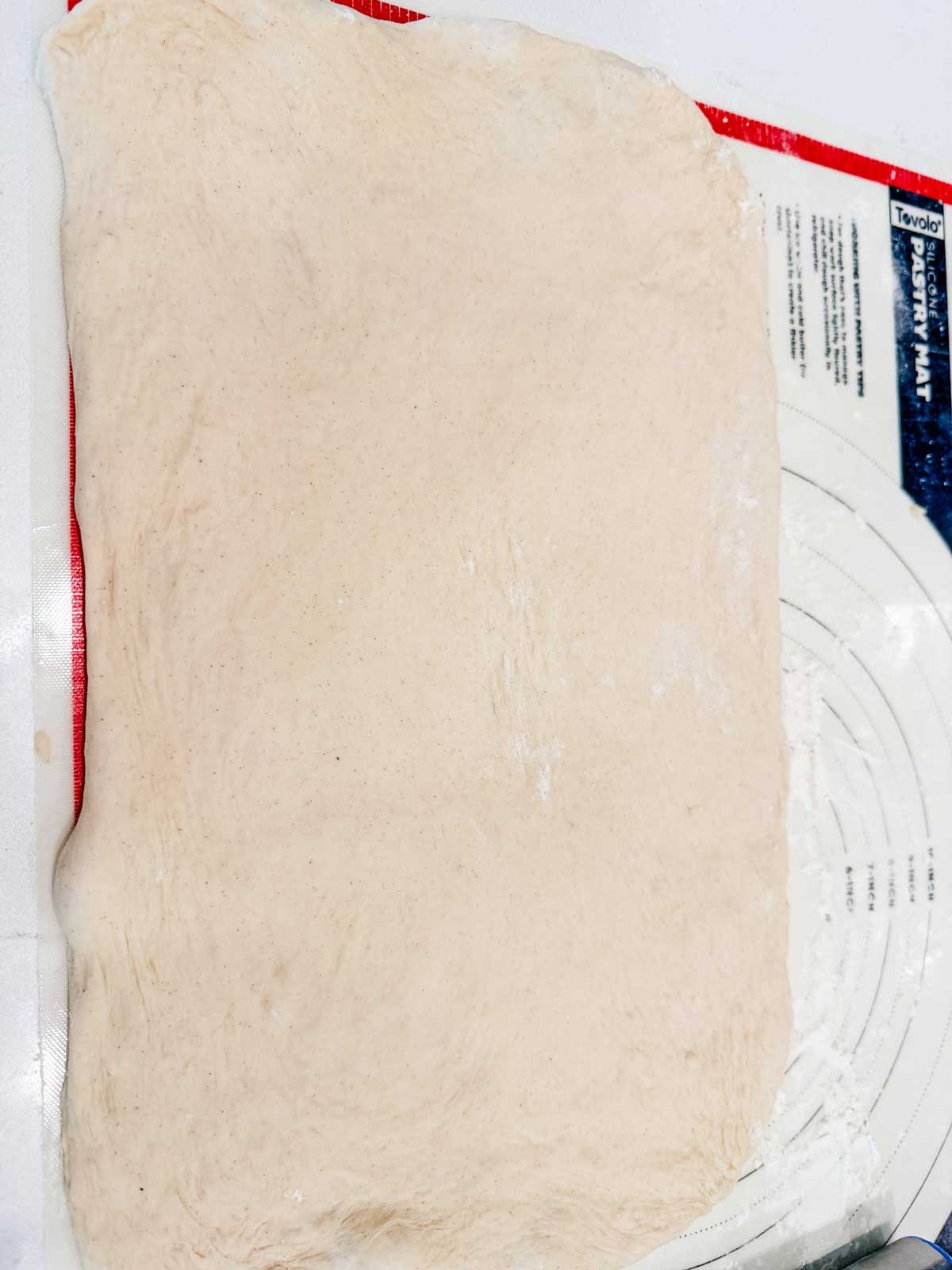 A rectangular sheet of raw, stretched dough rests on a silicone baking mat with measurement markings visible along the edge.