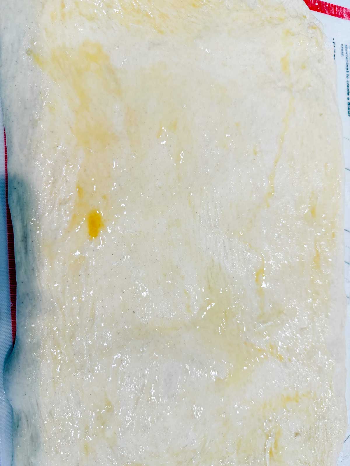 A close-up of stretched raw dough with a slightly glossy, oily surface, possibly prepared for baking or cooking, lying on a flat surface.