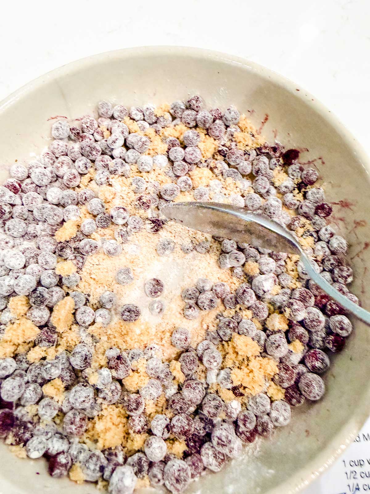 A bowl filled with blueberries, brown sugar, and flour being mixed with a metal spoon, viewed from above on a light surface.