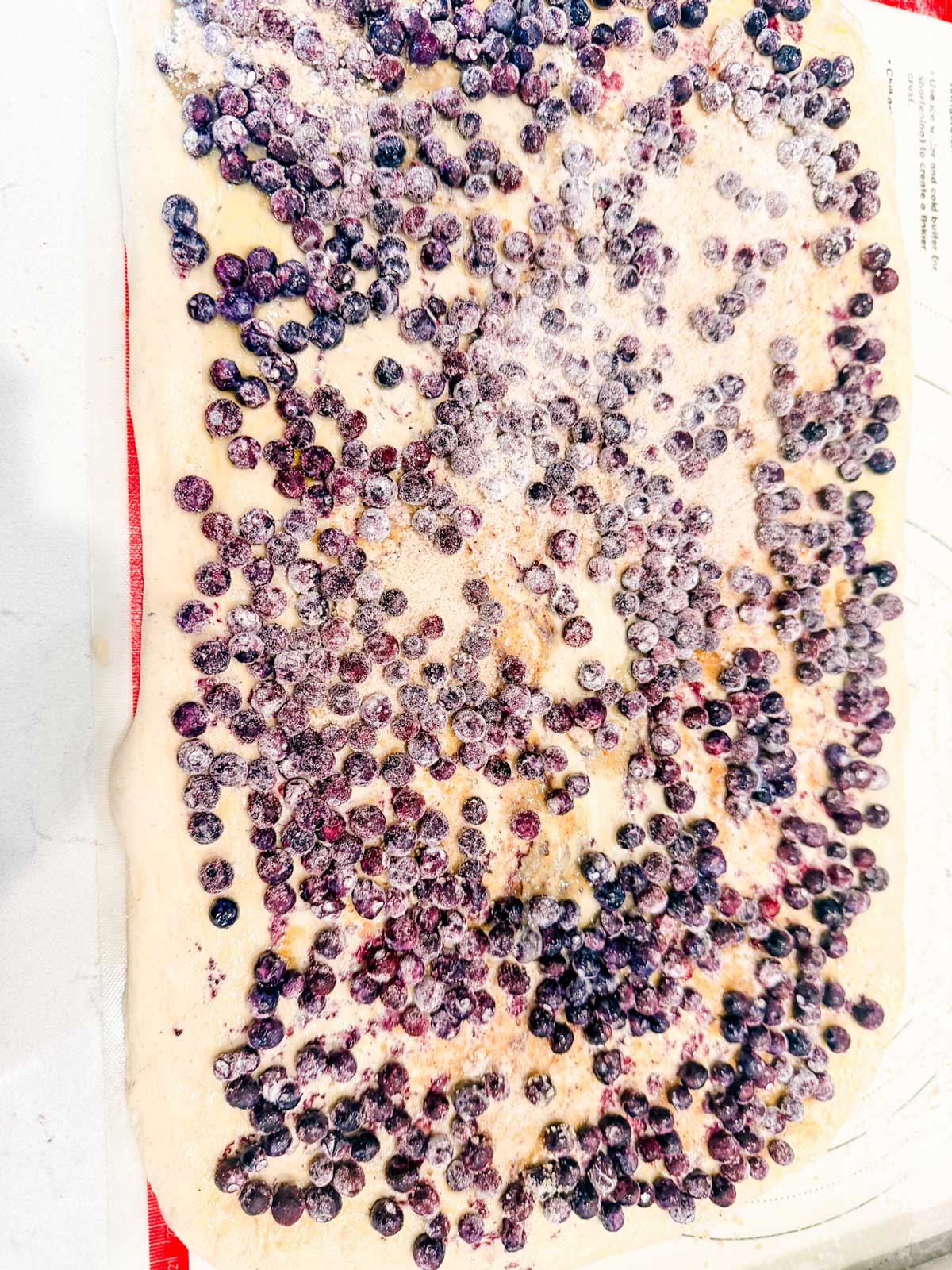 A sheet of dough is spread out on a surface and topped with a layer of frozen blueberries and a dusting of sugar.