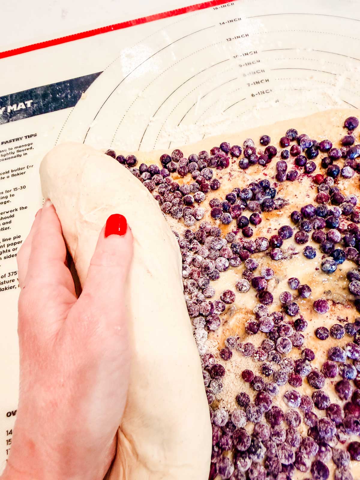 A hand with red nail polish rolls up dough covered with blueberries on a lightly floured pastry mat with measurement guides.