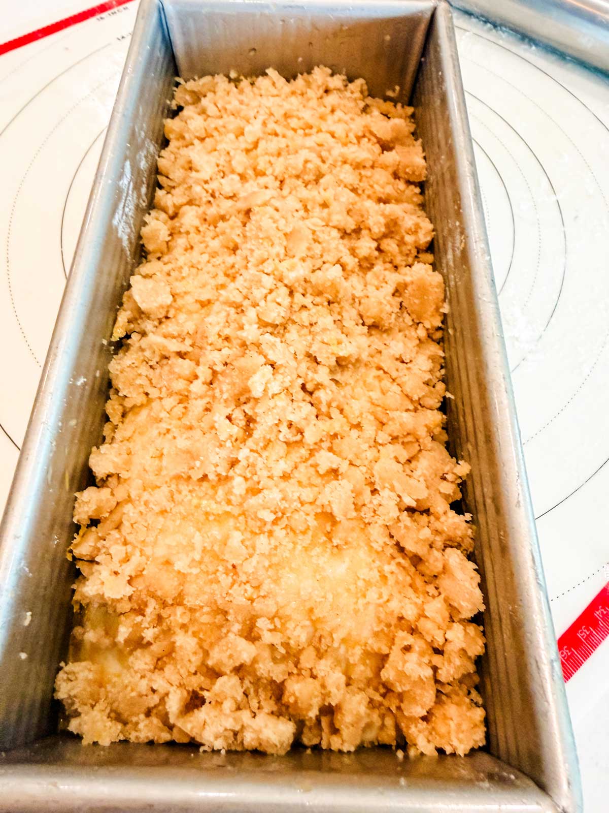 A close-up of an unbaked loaf in a metal pan, topped with a layer of crumbly streusel, sitting on a circular-patterned baking mat.
