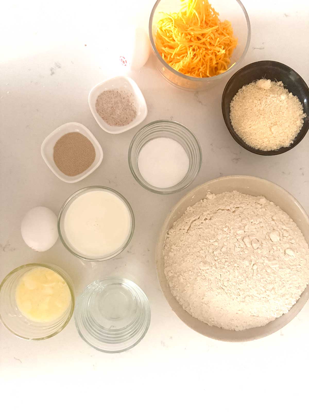 Overhead view of baking ingredients in bowls, including shredded cheese, flour, grated cheese, yeast, salt, sugar, milk, melted butter, water, and an egg, arranged on a light countertop.
