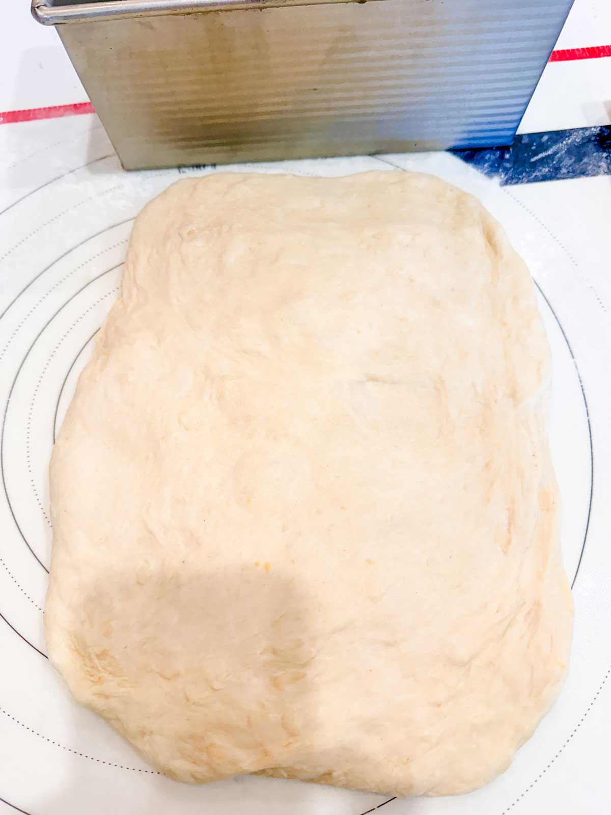 Rectangular piece of raw bread dough is flattened on a white surface with faint circular lines, next to a metal loaf pan.
