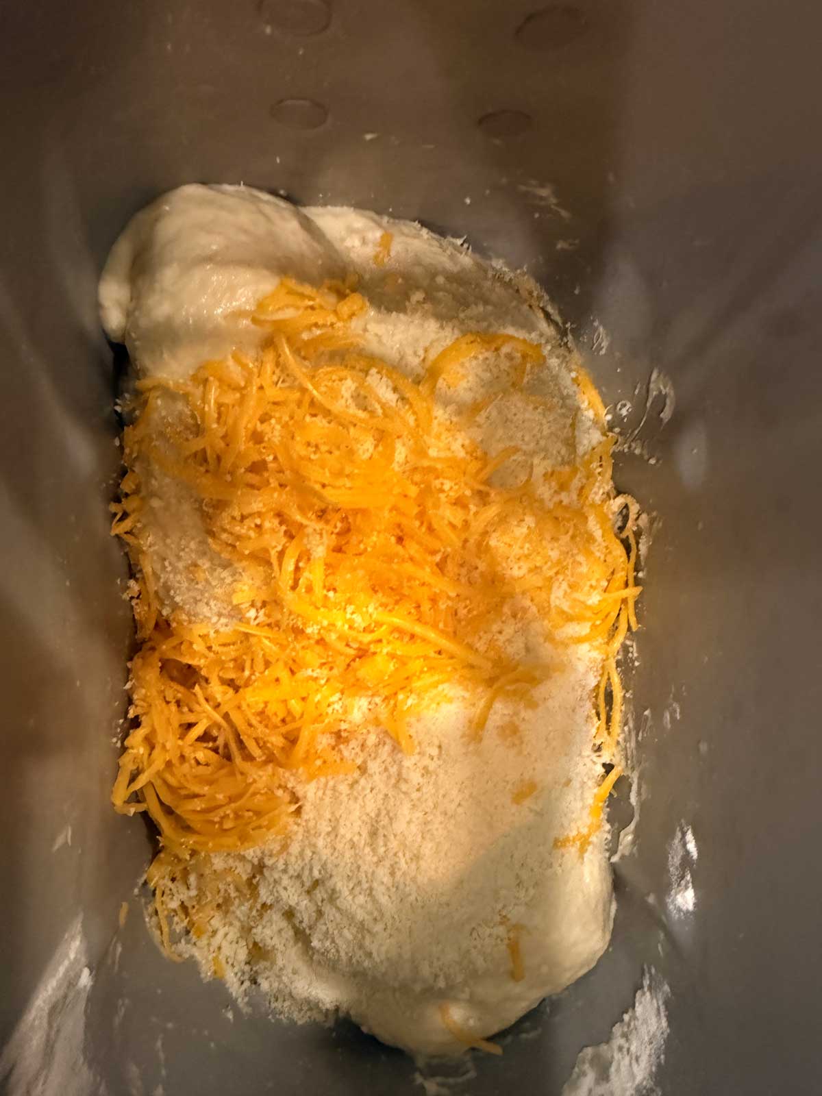 A close-up of shredded cheddar cheese and grated parmesan on top of dough inside a bread machine pan, ready to be mixed.
