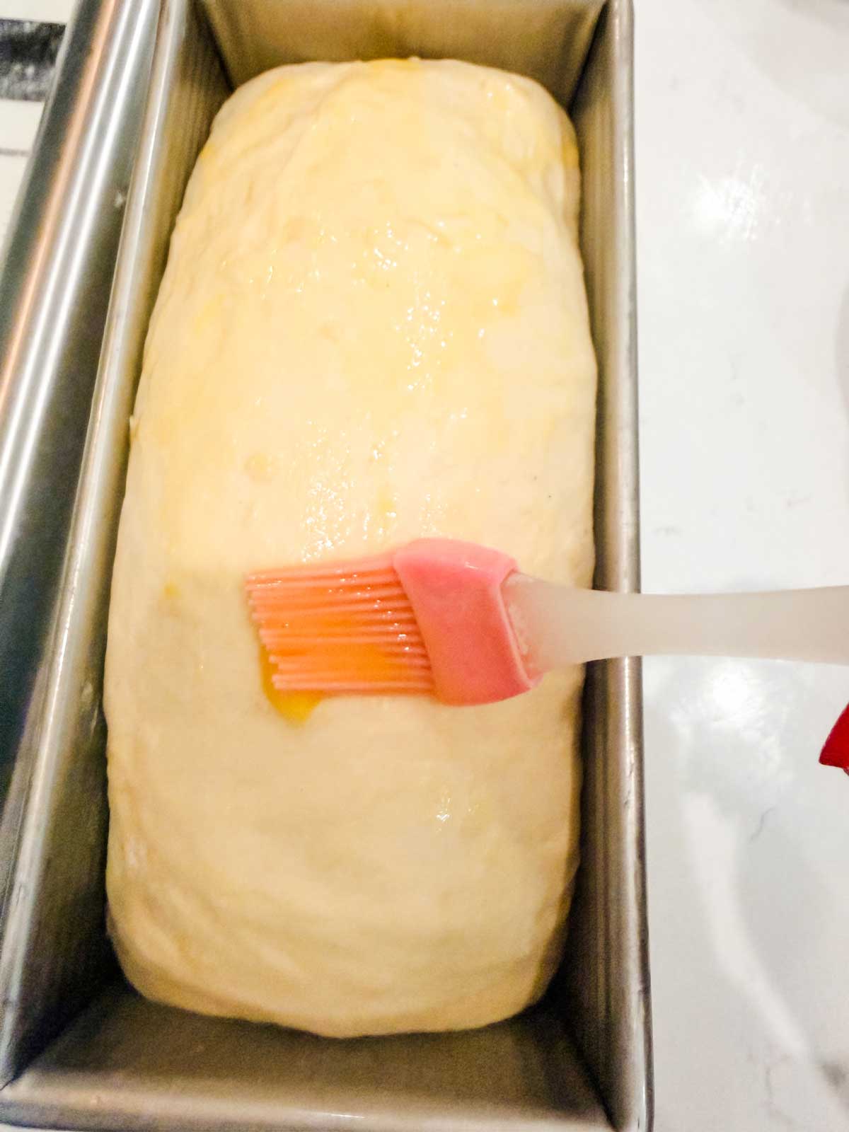 A loaf of unbaked bread dough in a metal pan being brushed with a glaze using a pink silicone pastry brush.
