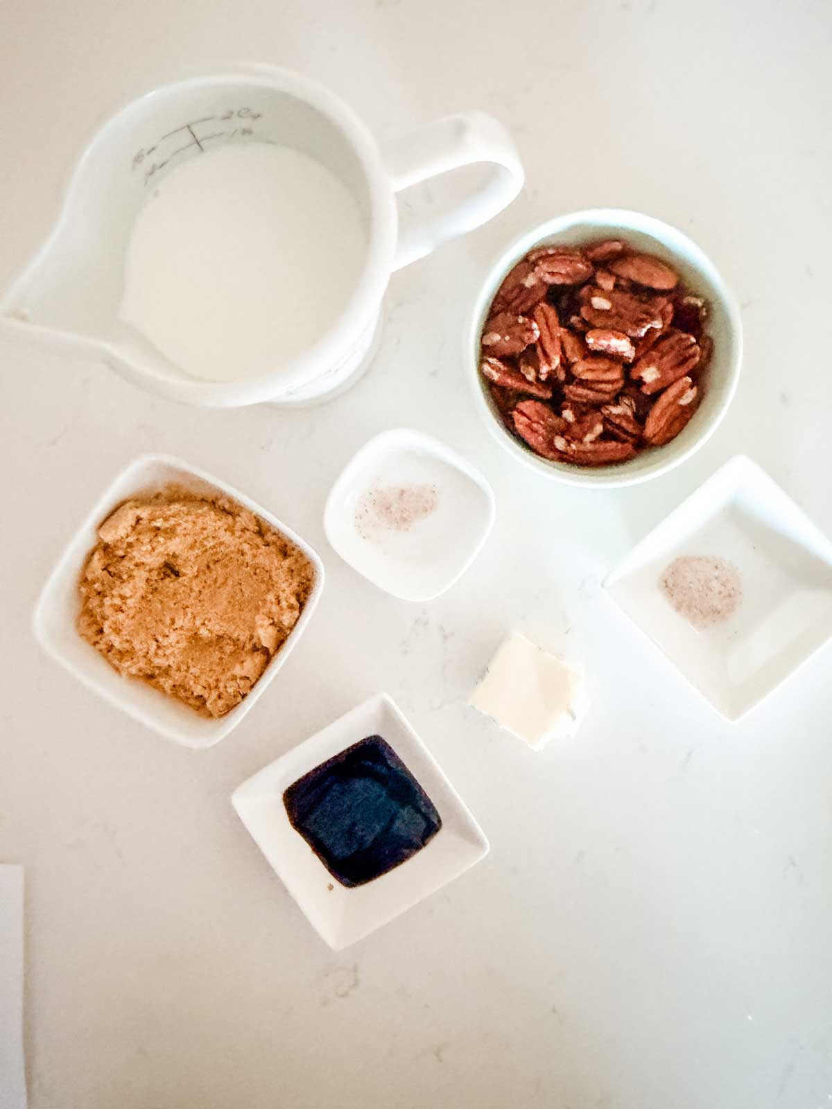 A top-down view of various ingredients in small bowls: milk, pecans, brown sugar, salt, butter, vanilla extract, and another small dish with a light powder, all arranged on a white surface.